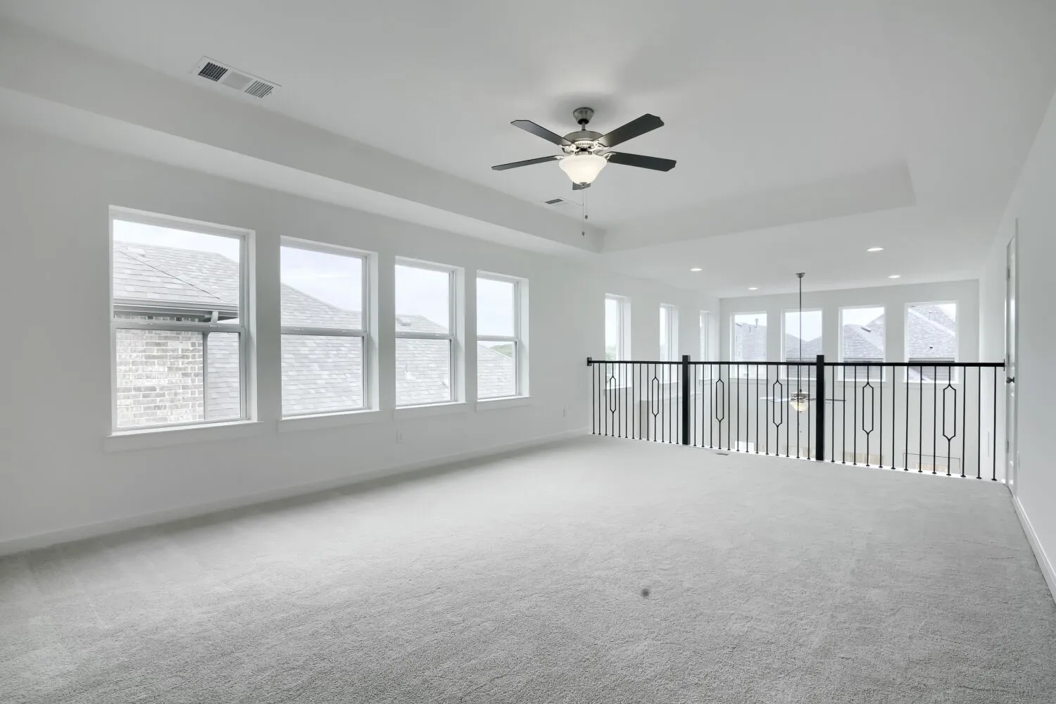 Spacious room featuring light gray carpeting, multiple windows, a ceiling fan, and a balcony with black metal balusters