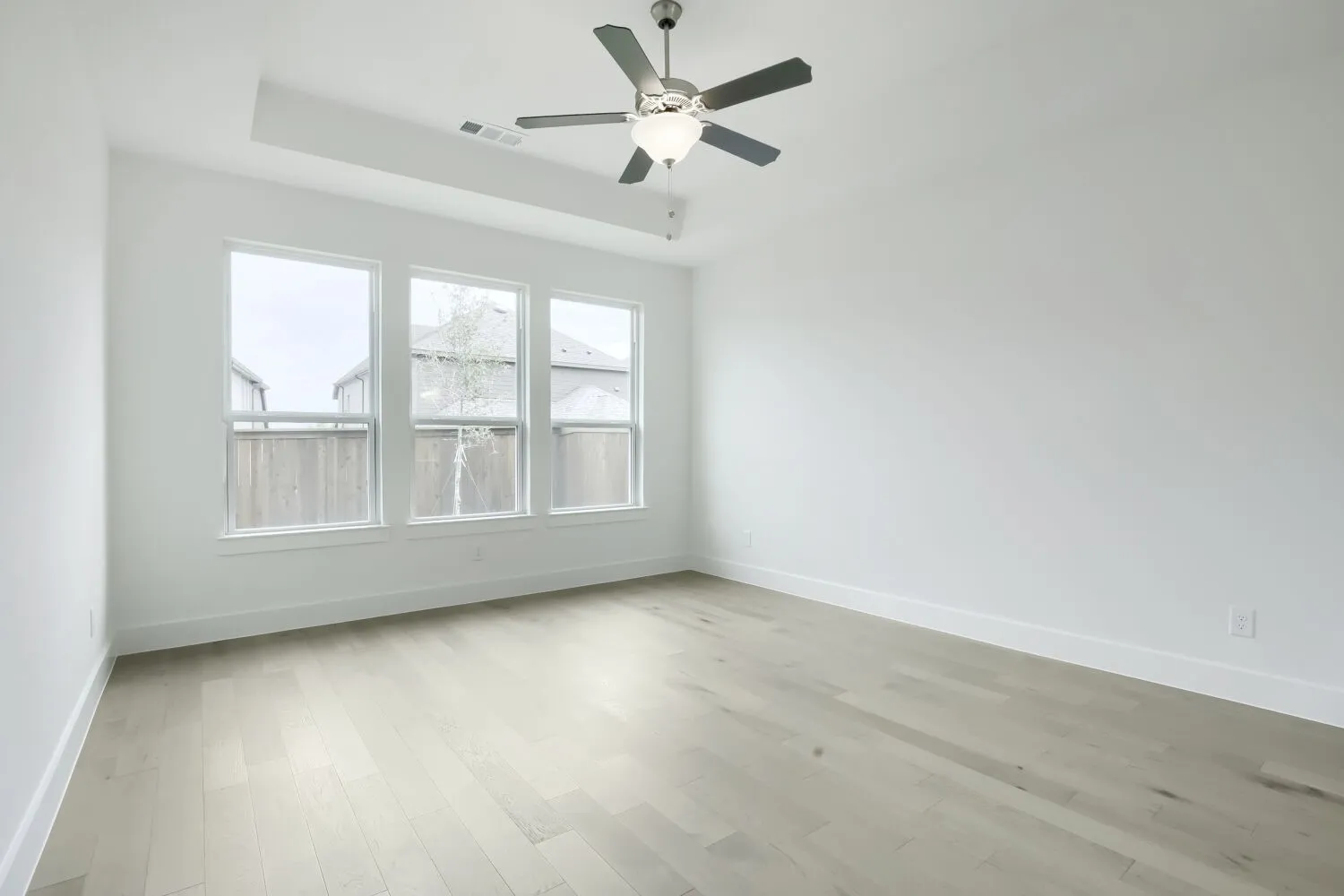 Spacious room featuring three large windows, light wood-finish flooring, a contemporary ceiling fan with integrated lighting, a tray ceiling detail, and crisp white walls with matching baseboards