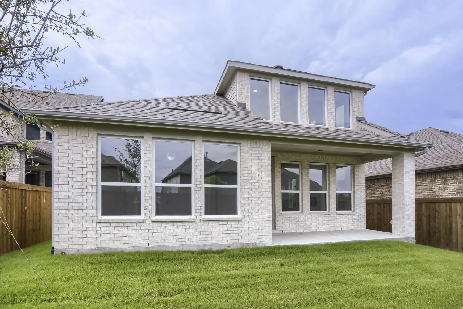 Rear exterior featuring an expansive green lawn, light-colored brick facade, covered patio, and multiple large windows