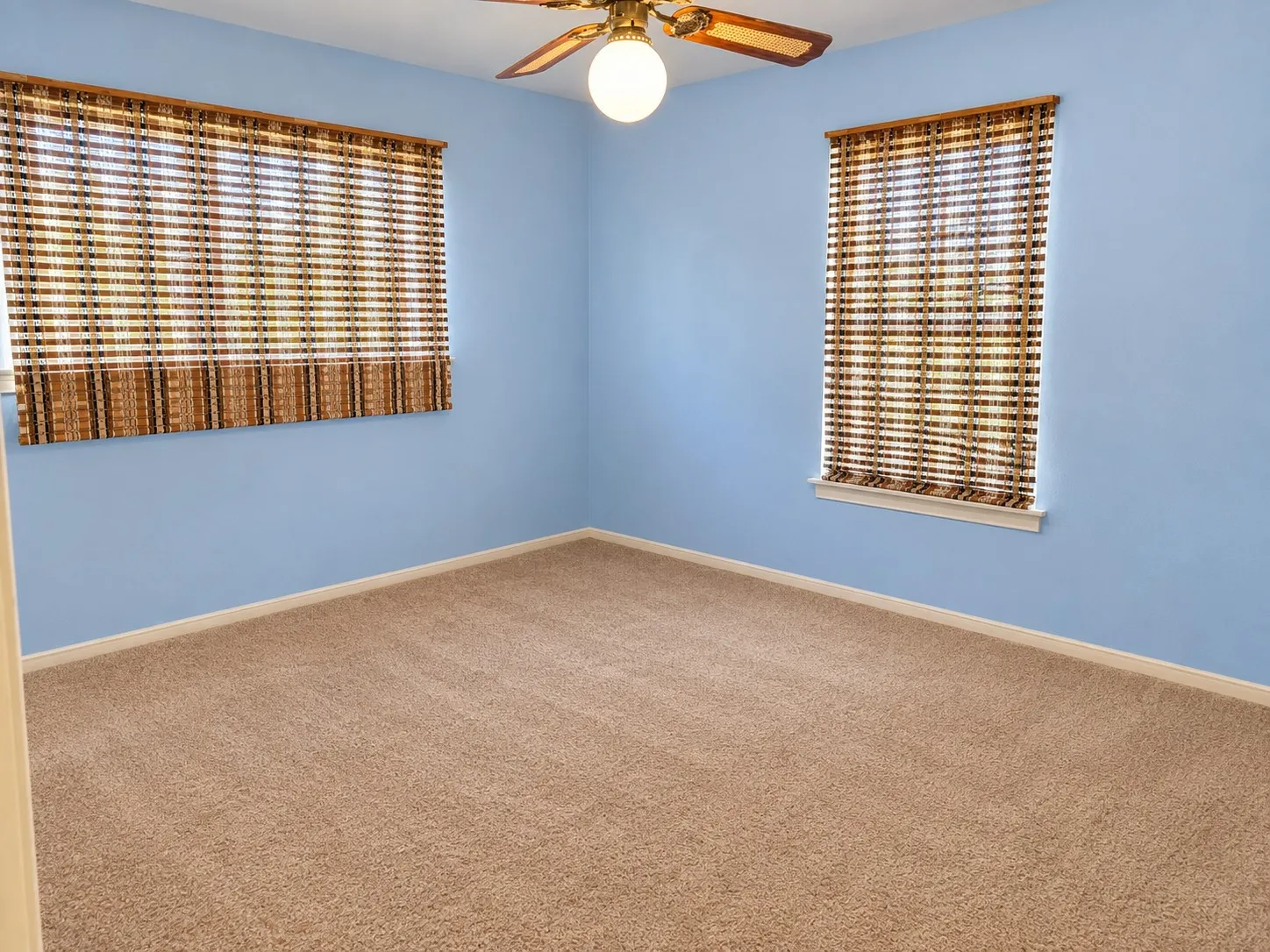 Textured carpet flooring, two windows with woven blinds, a ceiling-mounted light fixture, and a ceiling fan