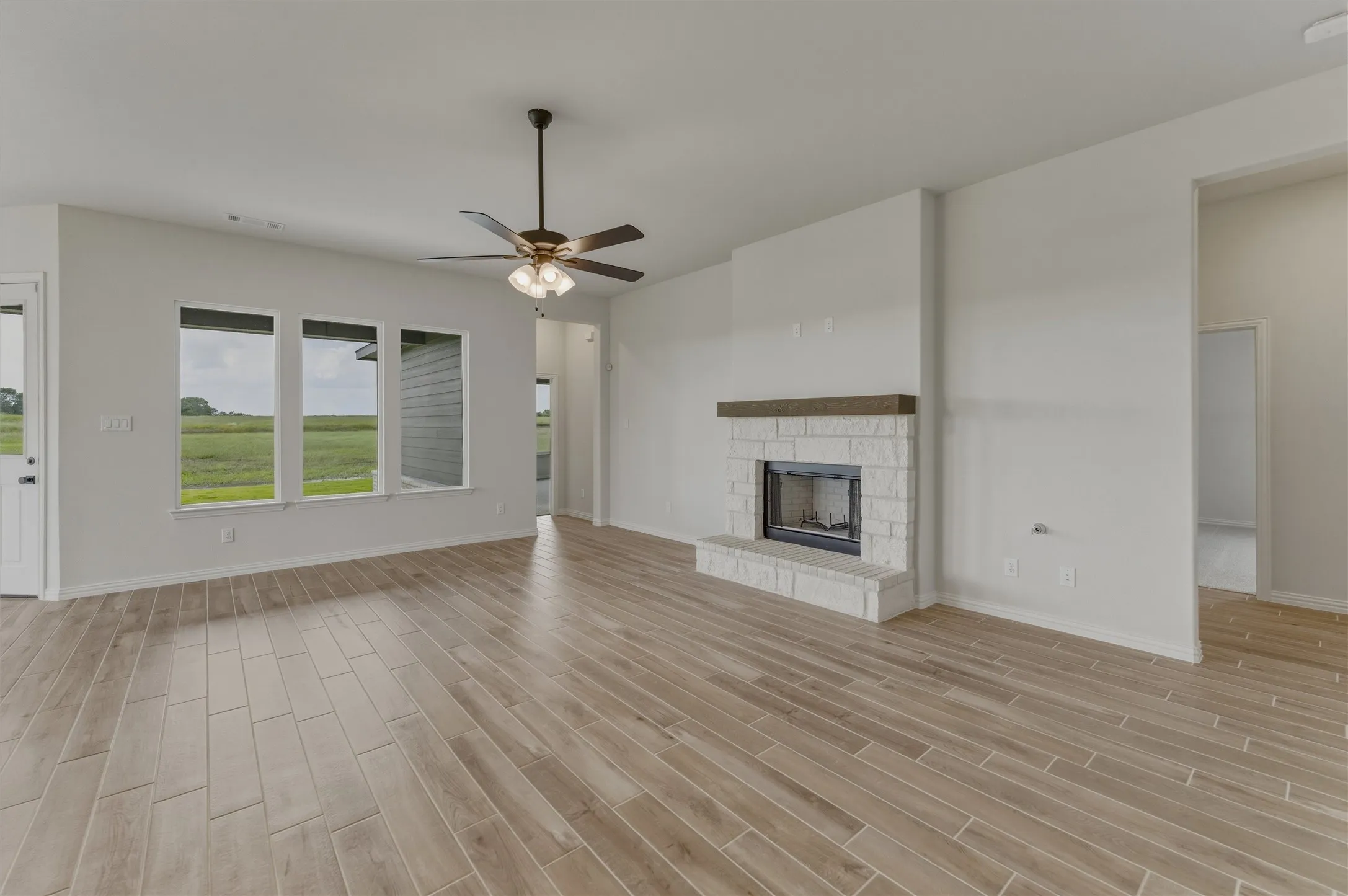 Spacious living area featuring wood-finish tile flooring, a stone-clad fireplace with a wood mantle, and three large windows providing ample natural light