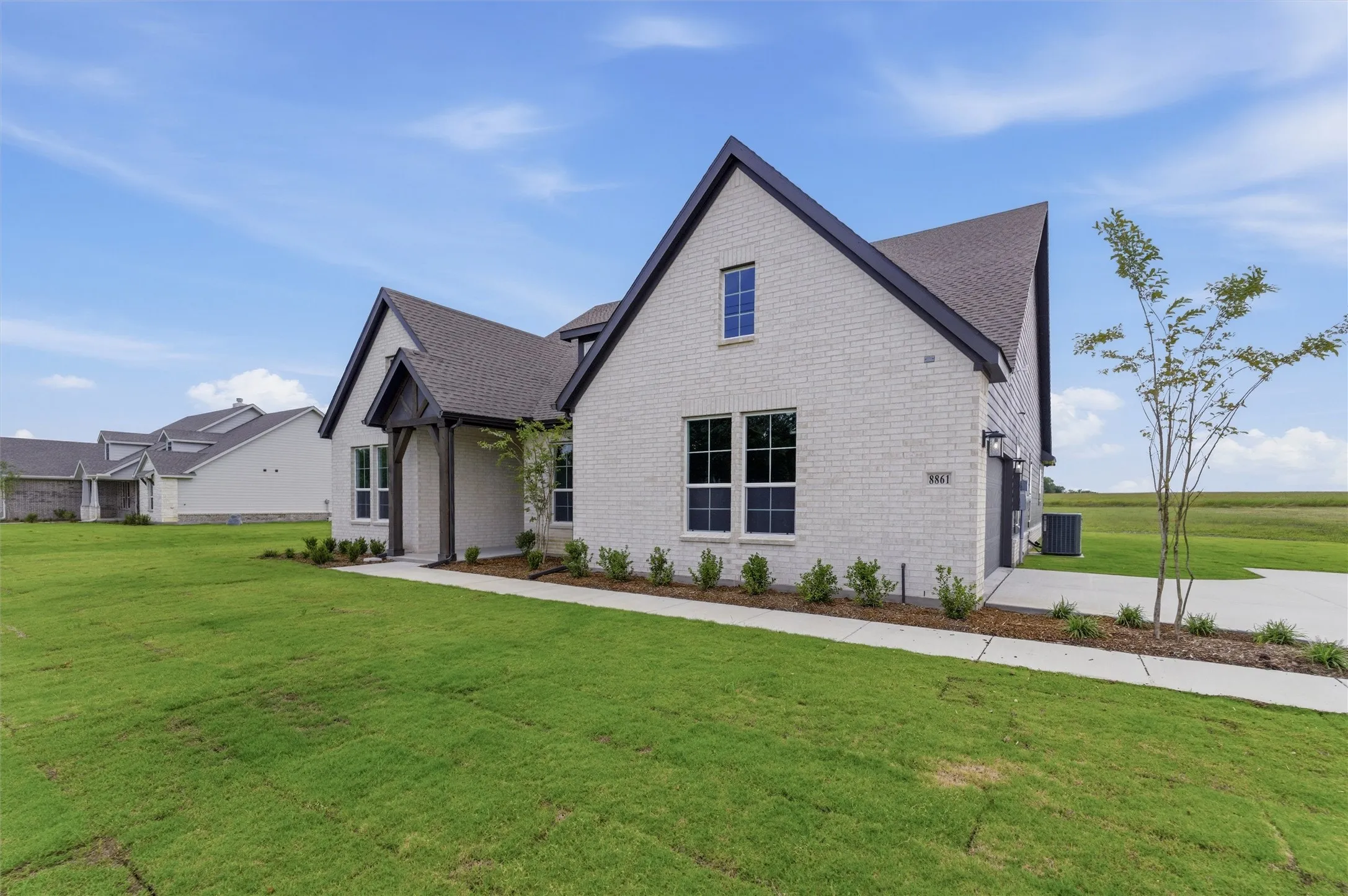 Modern farmhouse-style exterior featuring light-colored brick, dark-trimmed gables, a covered front entry with wood columns, a concrete driveway, and a manicured lawn