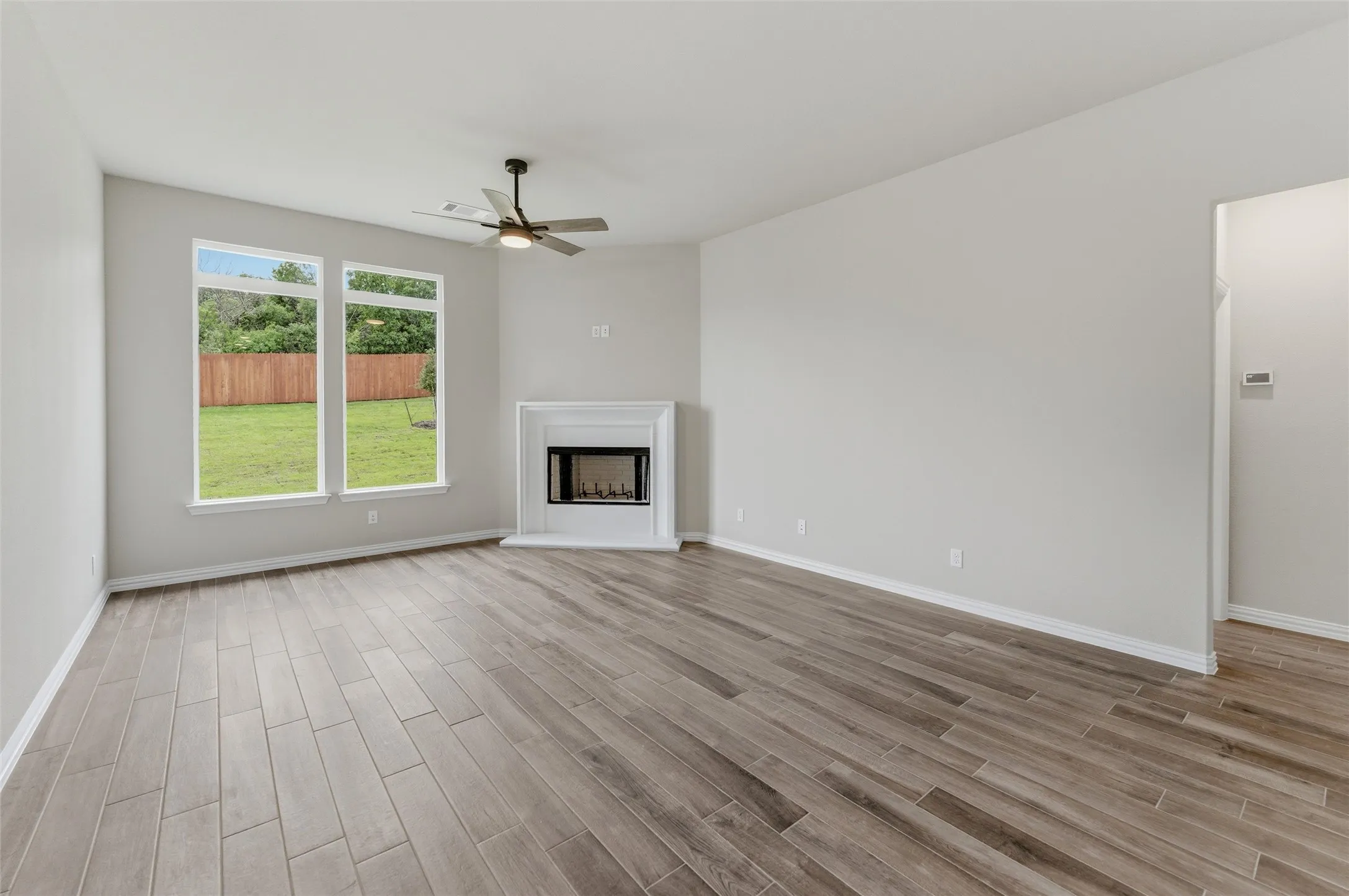 Spacious living area featuring wood-finish flooring, a corner fireplace with a white mantel, and two large windows overlooking a green lawn with a privacy fence