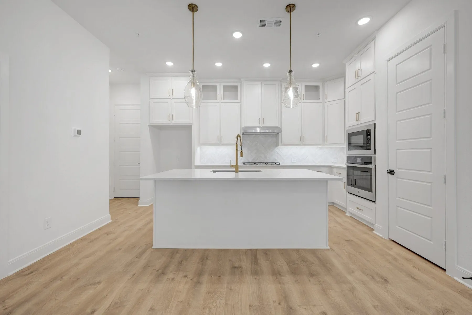 Open-concept kitchen featuring an expansive central island with a solid surface countertop, wood-finish flooring, white cabinetry, integrated stainless steel appliances, and two pendant light fixtures
