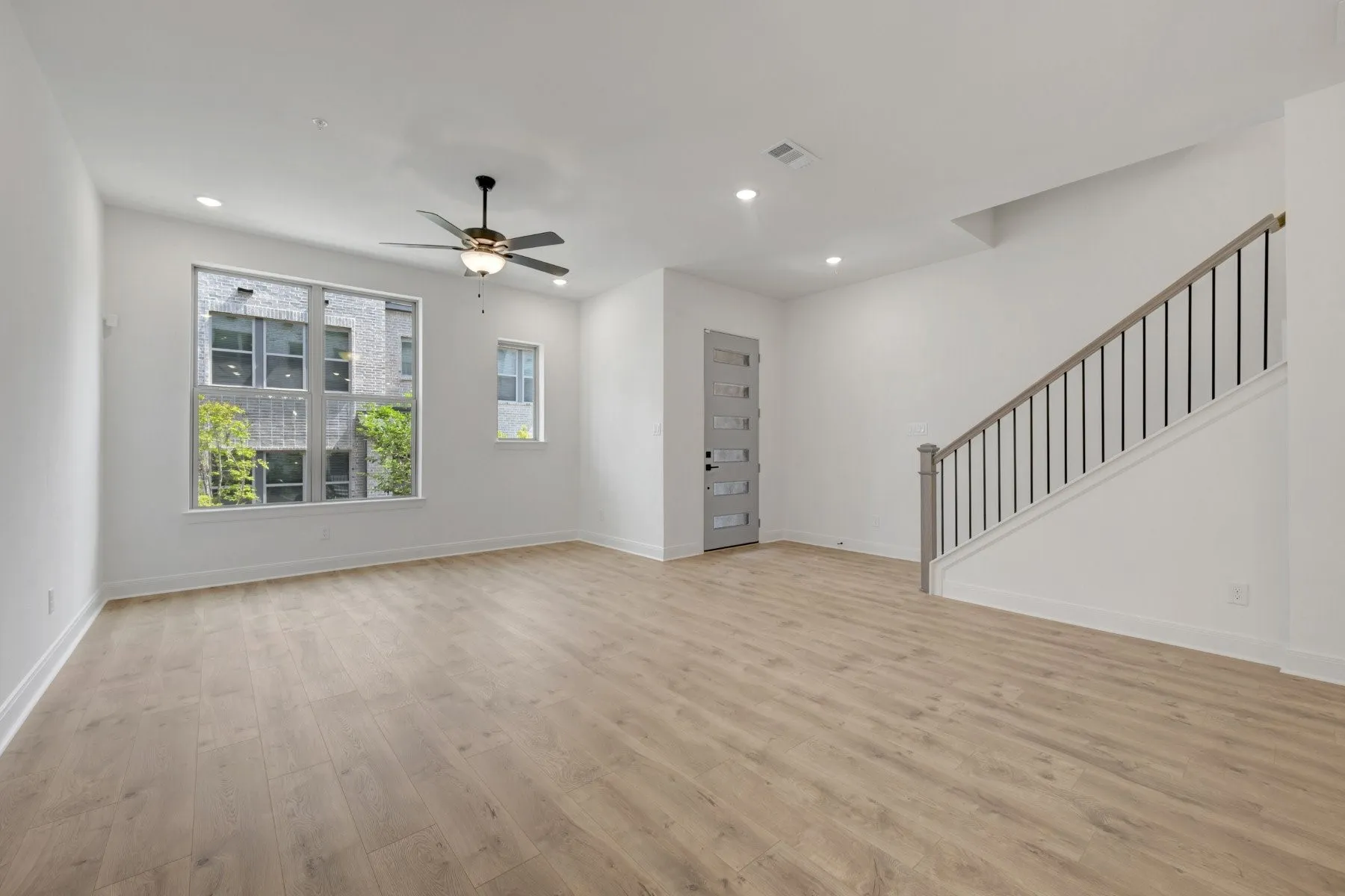 Spacious room featuring wood-finish flooring, a modern ceiling fan with lighting, recessed ceiling lights, and a prominent staircase with metal balusters