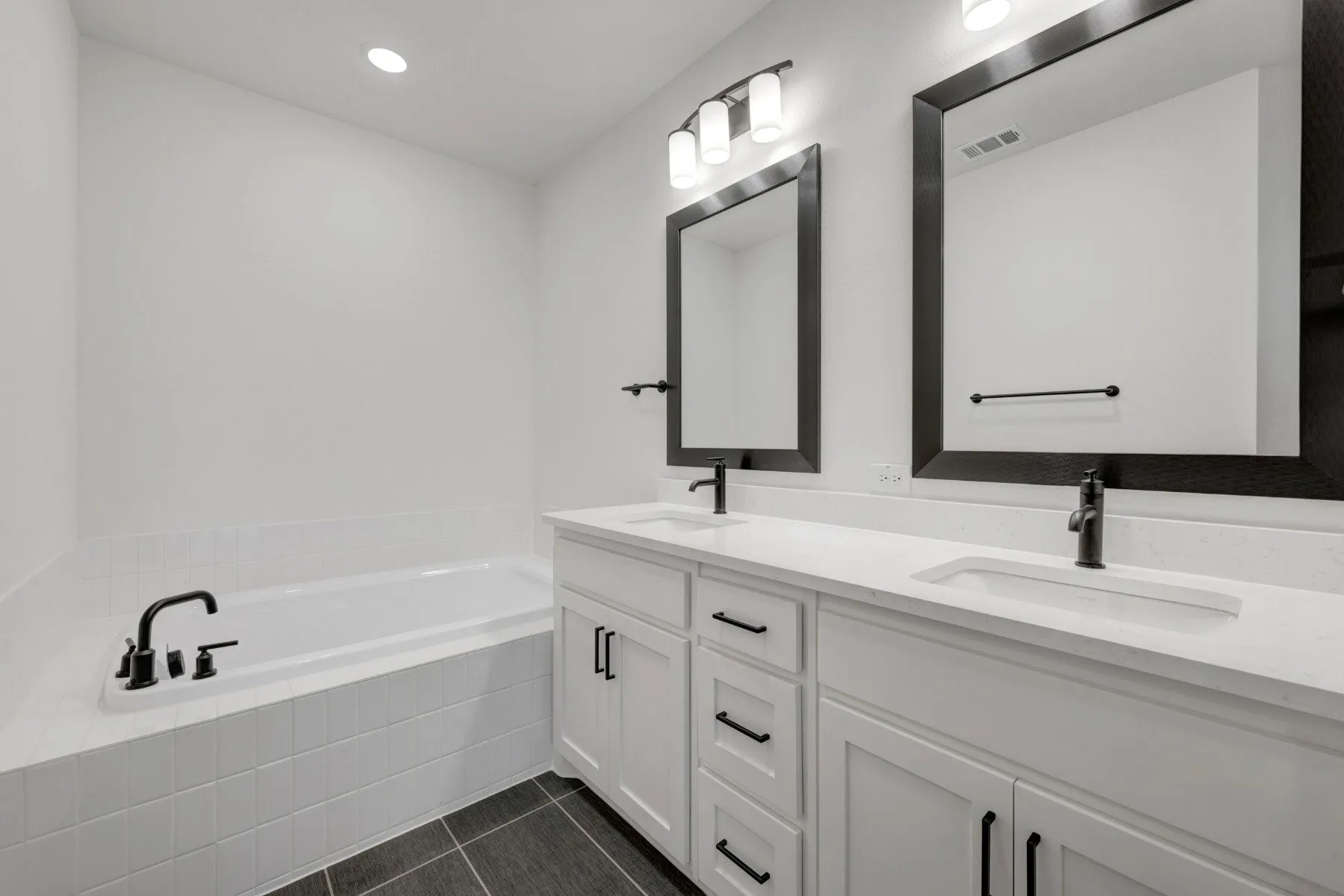 Bathroom featuring a dual vanity with white cabinetry and integrated sinks, matte black hardware, framed mirrors, and an inset soaking tub with white subway tile surround