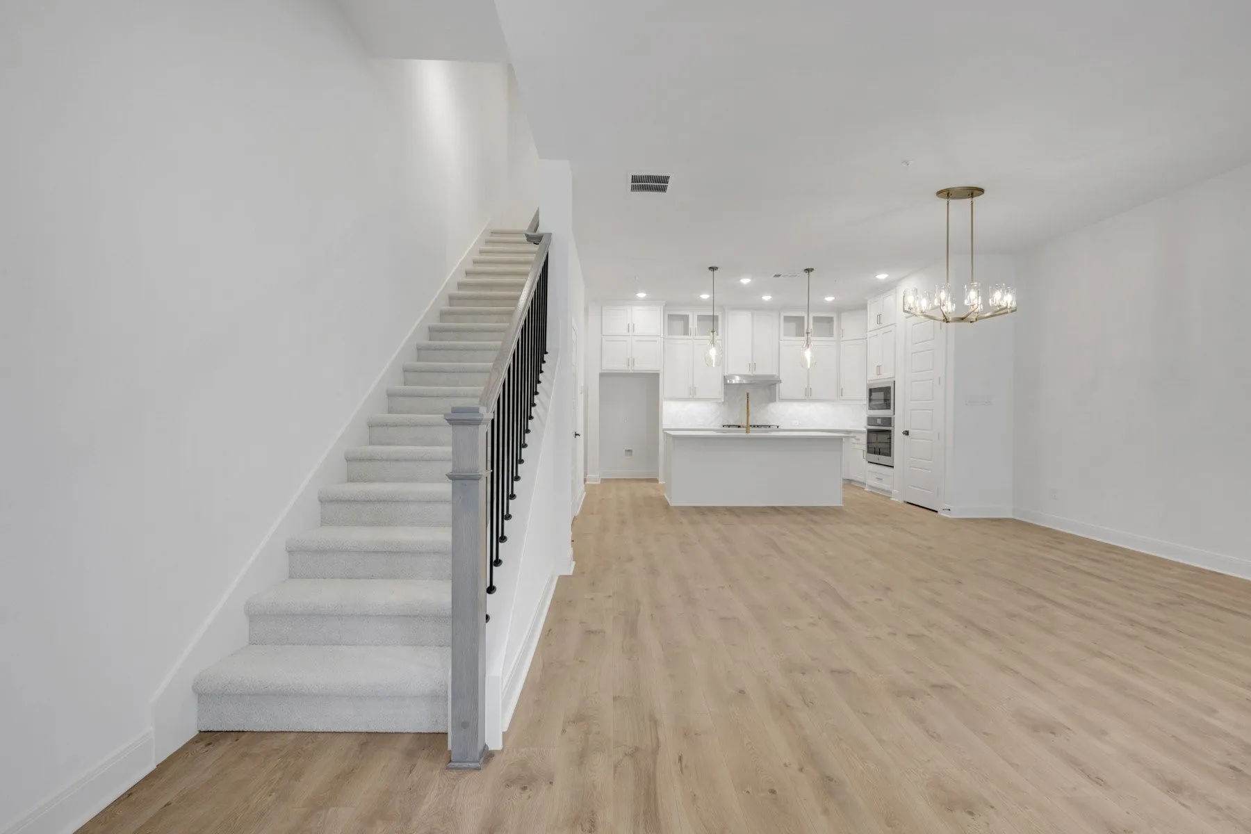 Open-concept living space featuring wood-finish flooring, a contemporary chandelier, and a carpeted staircase with a dark iron railing