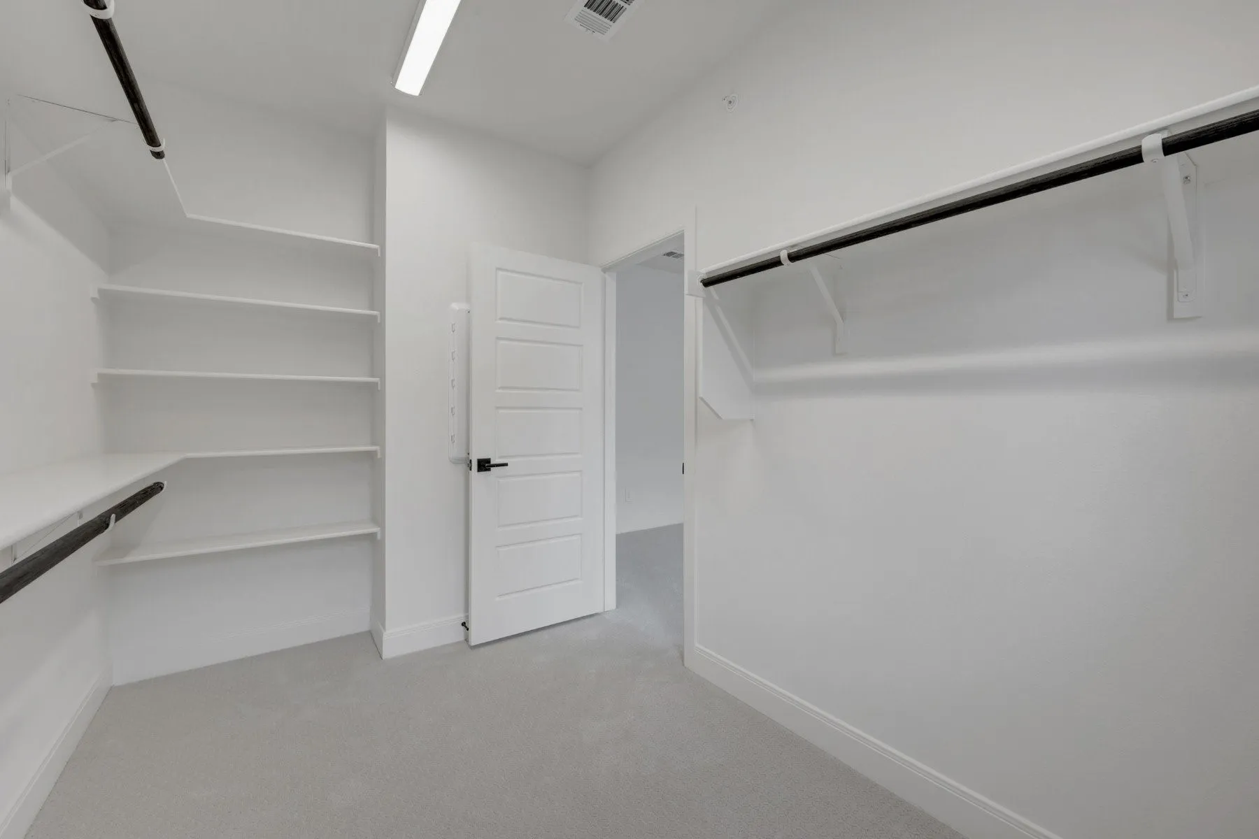 Spacious walk-in closet featuring extensive built-in shelving, dual hanging rods, light gray carpeting, a modern five-panel door with matte black hardware, and integrated overhead lighting