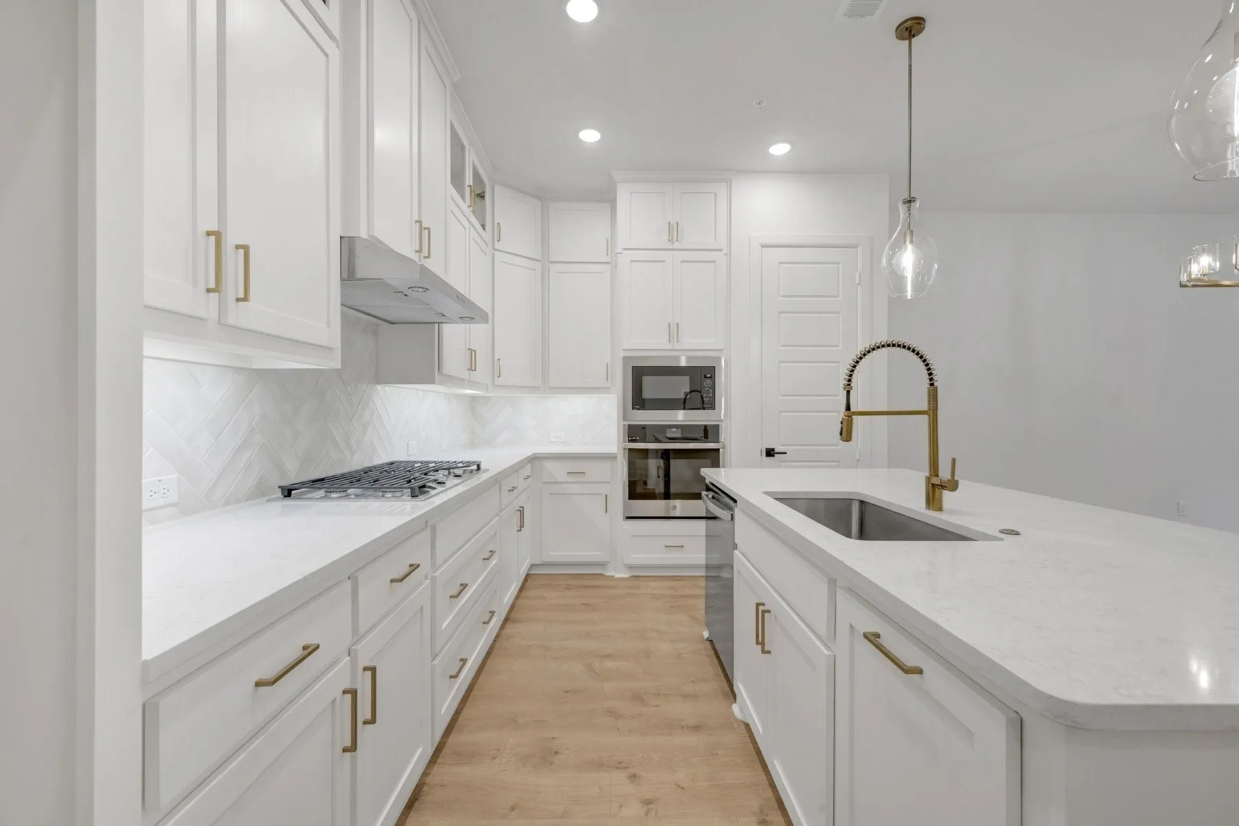 Kitchen featuring white shaker-style cabinetry with brass hardware, light-toned wood-finish flooring, white countertops, a herringbone tile backsplash, and stainless steel appliances