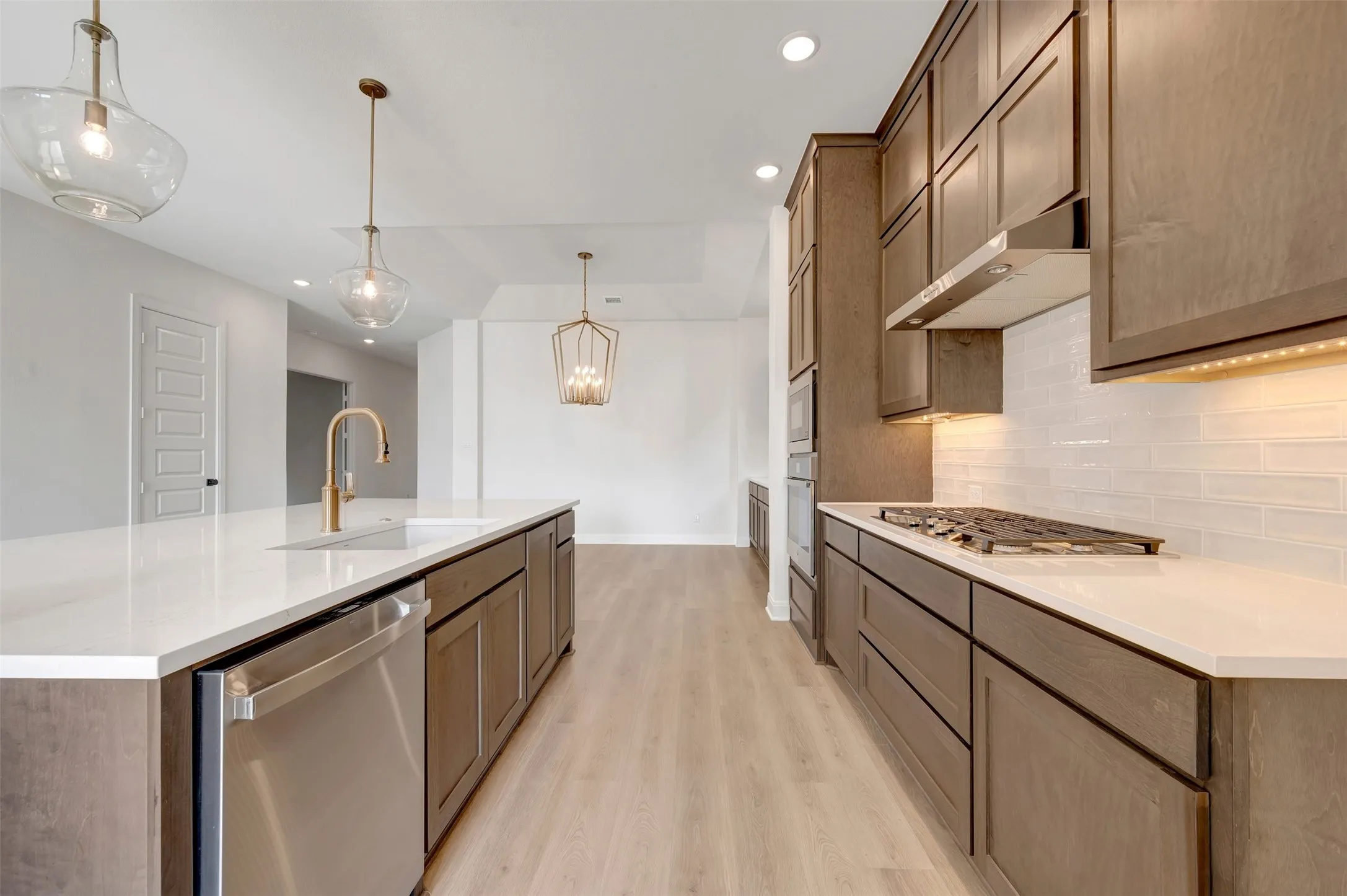 Contemporary kitchen featuring a spacious island with a gold-tone faucet, a stainless steel dishwasher, wood-finish cabinetry, a gas cooktop with a stainless steel range hood, and light wood-finish flooring