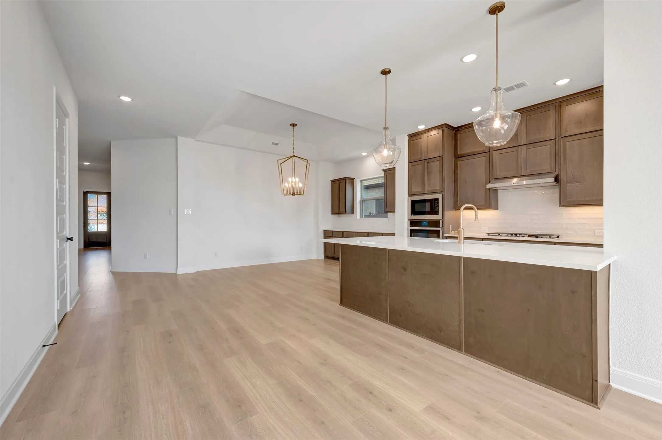 Open-concept kitchen and living area featuring light wood-finish flooring, a kitchen island with a white countertop, built-in stainless steel appliances, and suspended pendant lighting