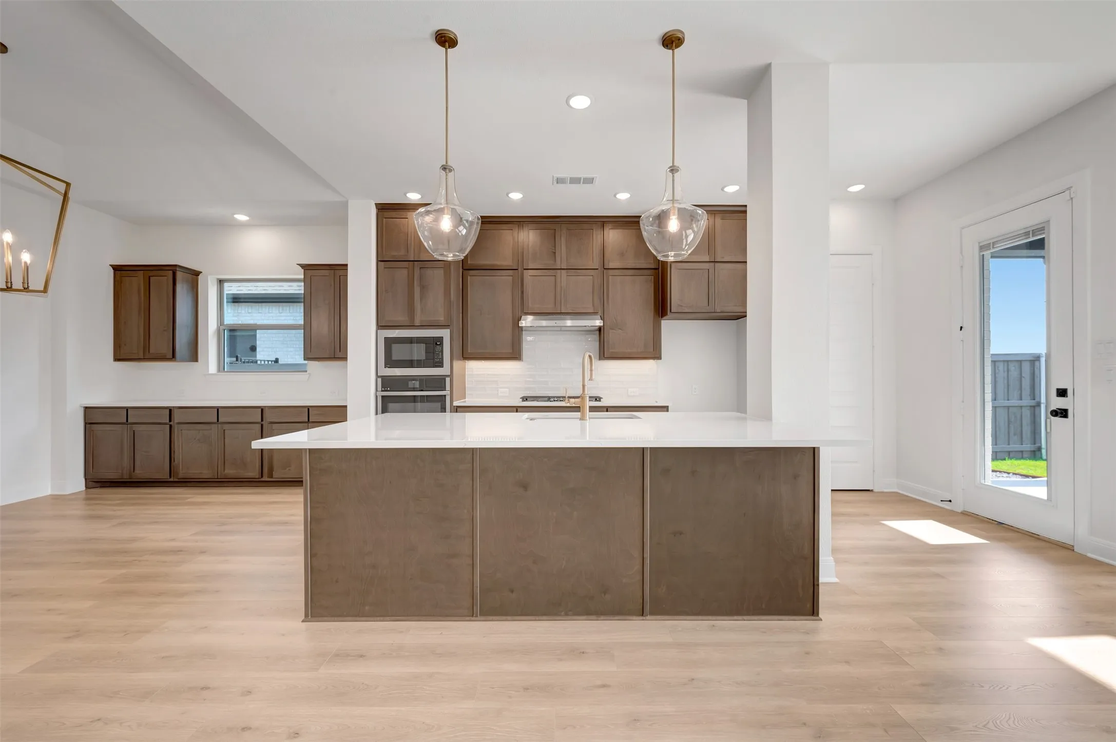 Open-concept kitchen featuring an oversized island with a white solid surface countertop, built-in stainless steel double ovens, wood-finish cabinetry, and light wood-finish flooring