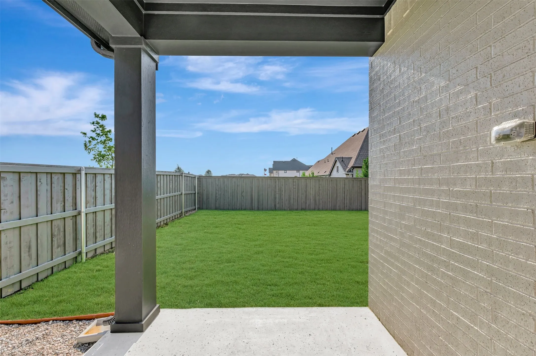 Covered patio with a concrete surface, featuring grey brick exterior, dark grey support columns, and a fully fenced backyard with green lawn