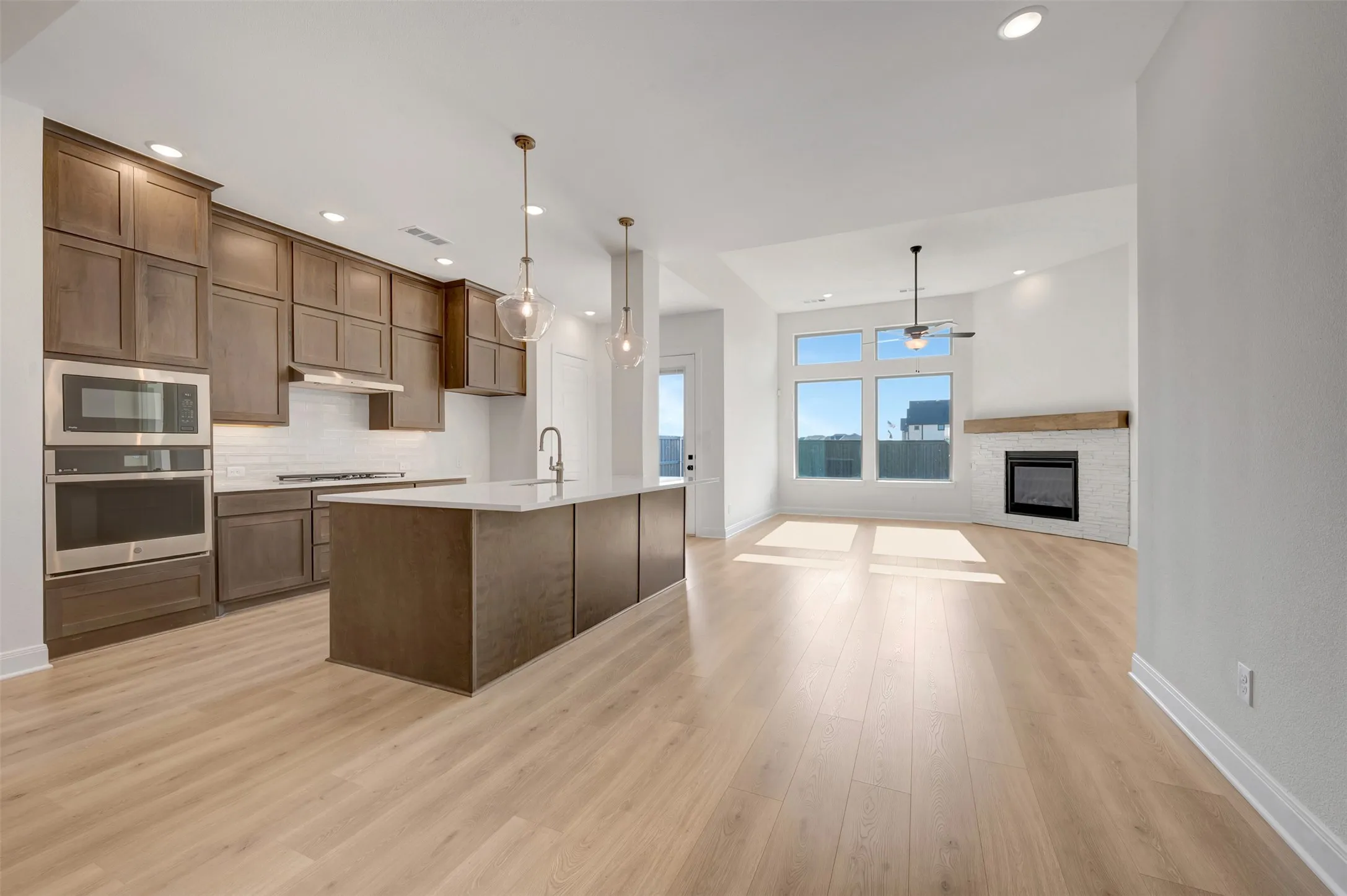 Open-concept living space with light wood-finish flooring, a kitchen island featuring a sink and pendant lighting, built-in stainless steel appliances, a tile-surround fireplace with a wood mantle, and large windows