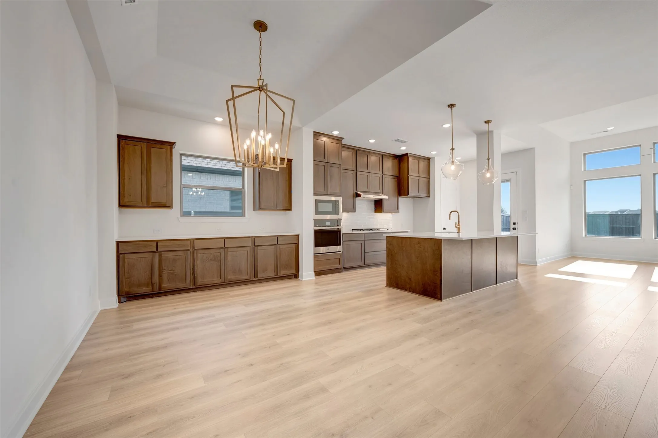 Open-concept living space with light wood-finish flooring, a kitchen island with pendant lighting, integrated stainless steel appliances, extensive cabinetry, and large windows