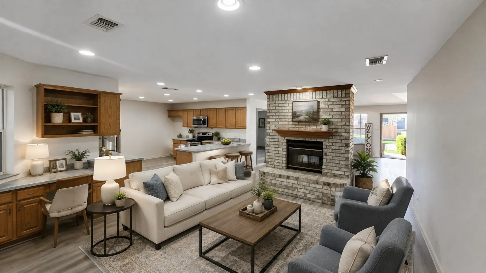 Virtually staged. Open-concept living space featuring a brick fireplace, built-in desk with cabinetry, wood-finish flooring, recessed lighting, and a kitchen with light wood cabinetry and stone countertops