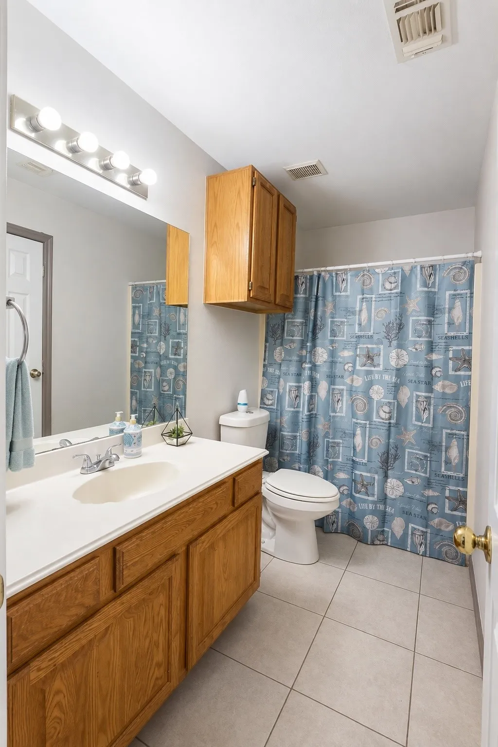 Bathroom featuring a wood-finish vanity with a white countertop and integrated sink, a wall-mounted mirror, overhead vanity lighting, and ceramic tile flooring