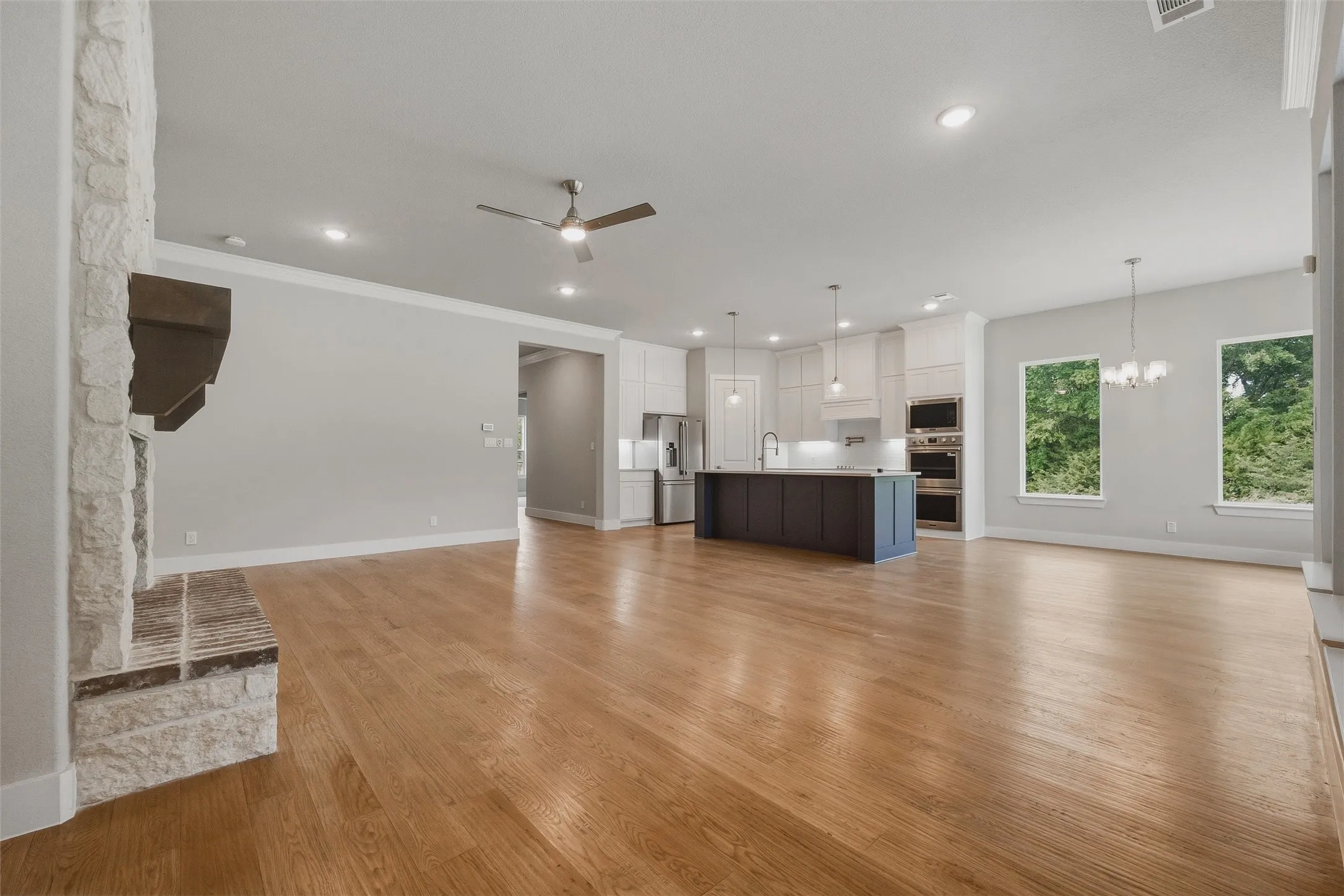 Stone-clad fireplace with wood mantel, wood-finish flooring, recessed lighting, and a kitchen featuring a dark-toned island with pendant lighting and stainless steel appliances
