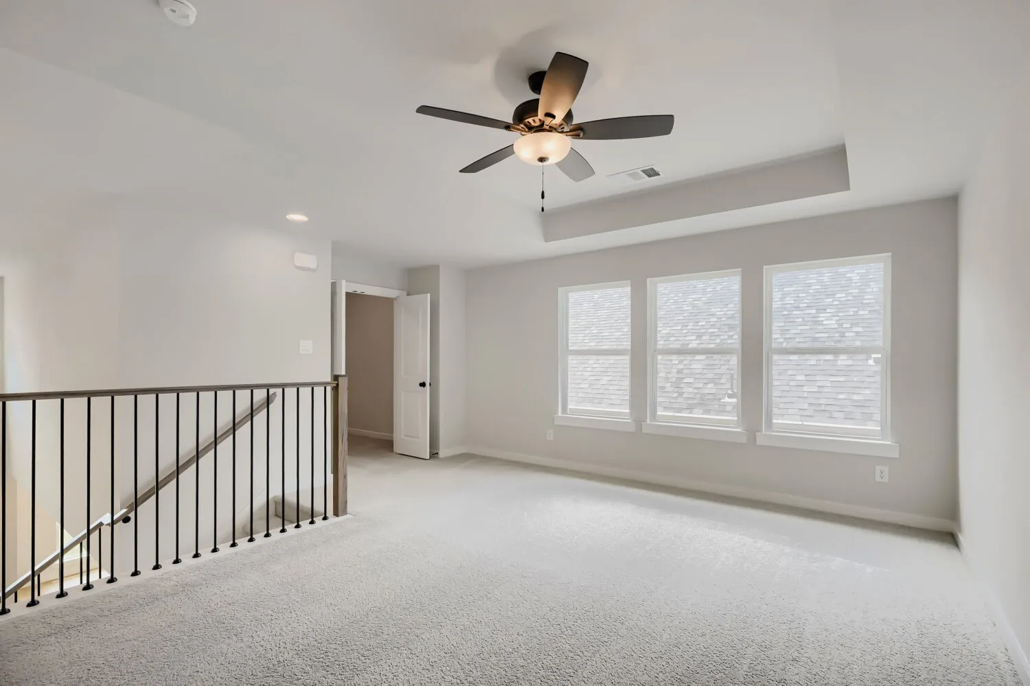 Loft space featuring light gray carpeting, three prominent windows, a ceiling fan with integrated lighting, and a tray ceiling detail