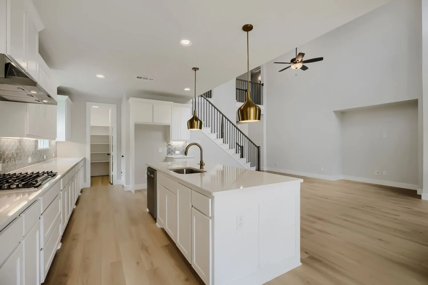 Open-concept living space featuring a kitchen island with sink and two brass pendant lights, stainless steel appliances, wood-finish flooring, and a two-story ceiling in the living area with a ceiling fan