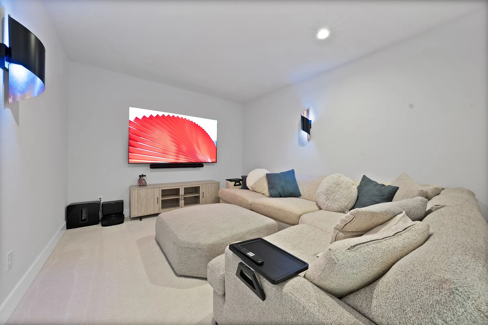 Carpeted entertainment area featuring white walls, a mounted television, two wall sconces, and a built-in media console with glass panel doors