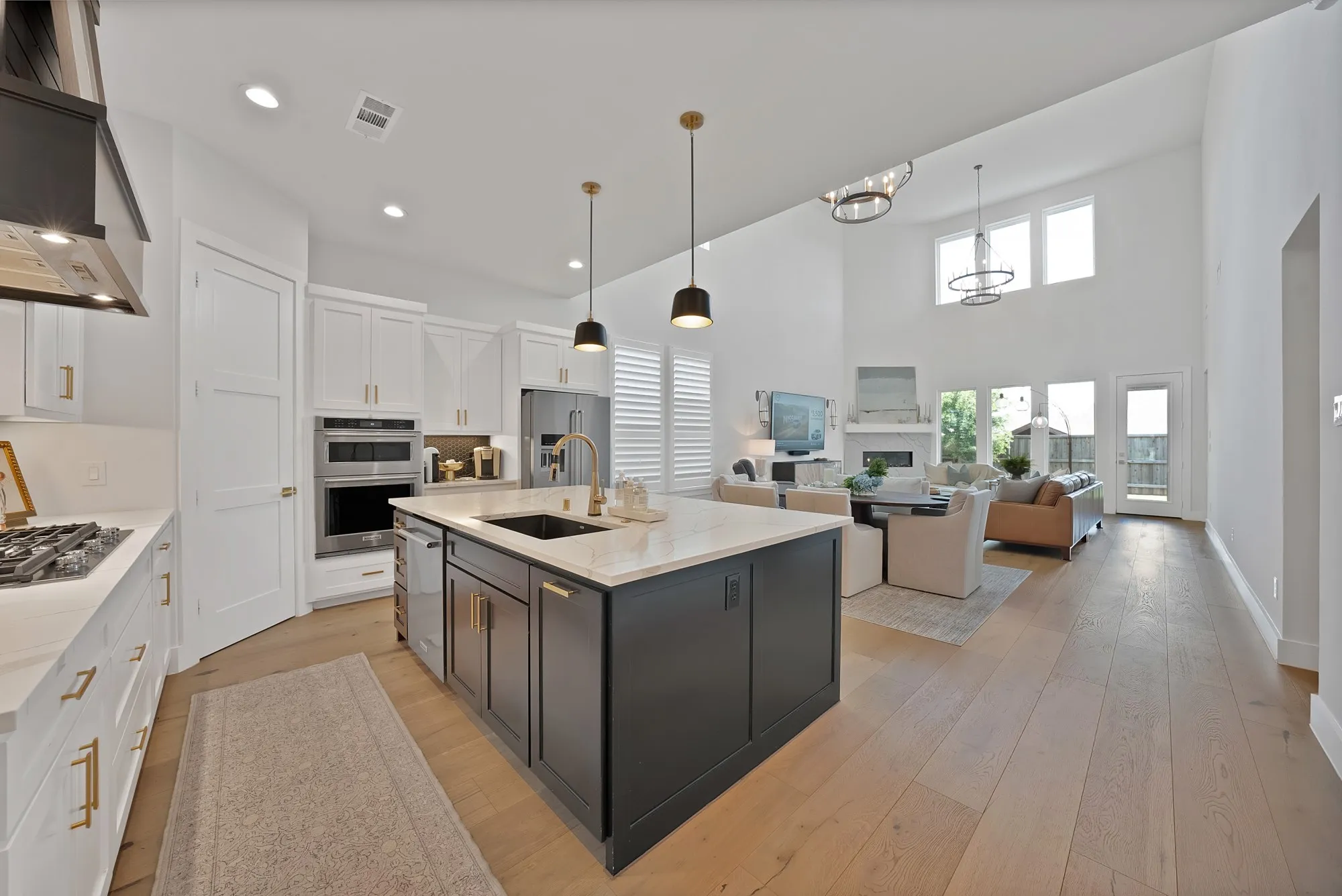 Spacious kitchen featuring an island with a sink, white cabinetry, integrated stainless steel appliances, and wood-finish flooring extending into a high-ceiling living area with large windows