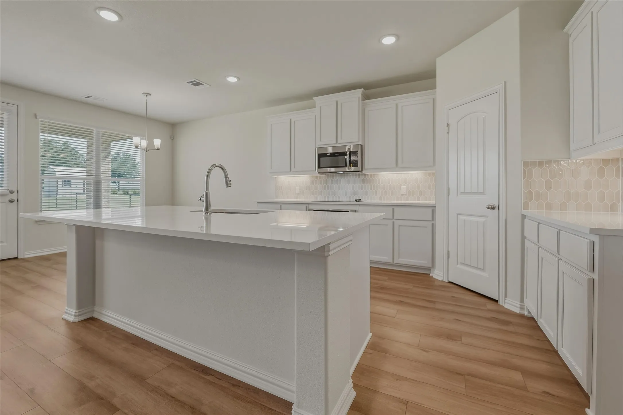 Bright kitchen featuring a center island with sink, stainless steel microwave, white cabinetry, hexagon tile backsplash, and wood-finish flooring