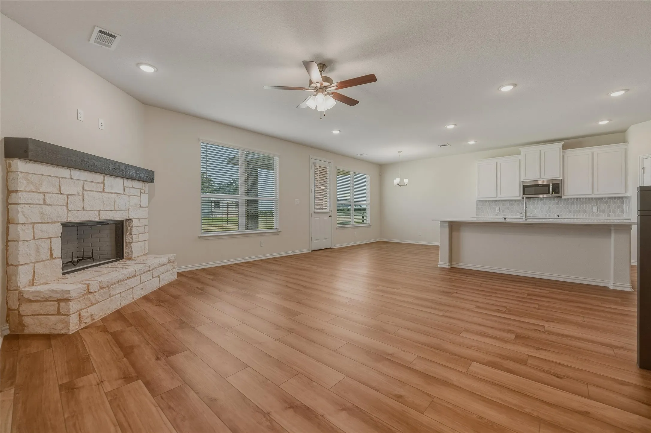 Expansive open-concept living area featuring wood-finish flooring, a stone-clad fireplace with a dark wood mantel, recessed lighting, and a ceiling fan