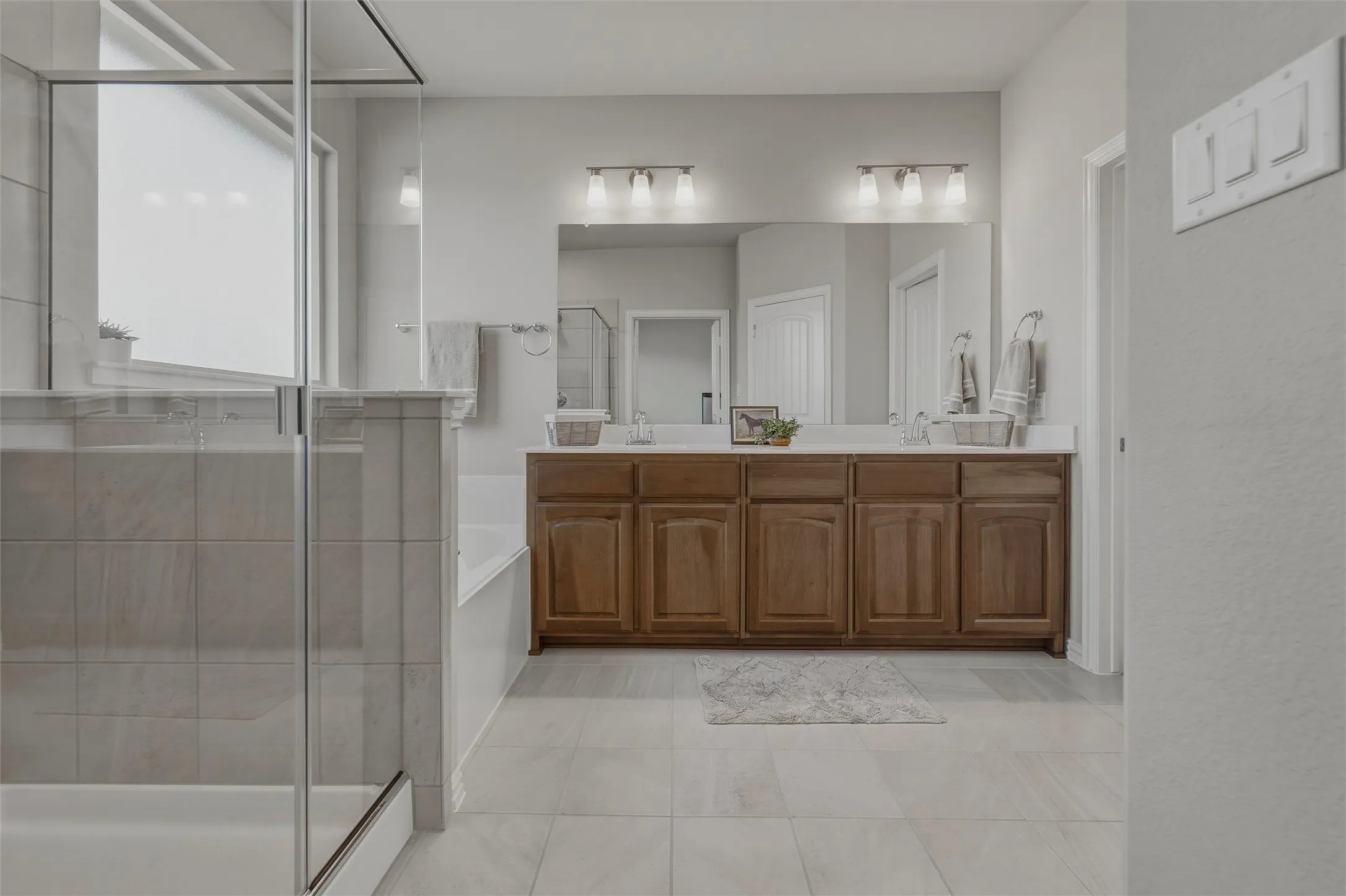 Dual vanity with wood-finish cabinetry and integrated sinks, large wall-mounted mirror, ceramic tile flooring, and glass-enclosed shower with tiled walls