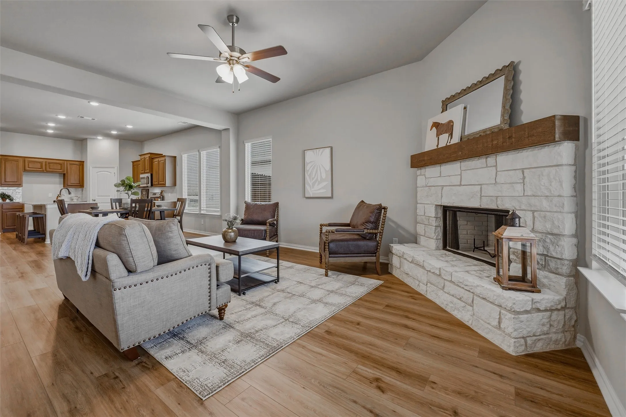 Spacious living area featuring wood-finish flooring, a stone-clad fireplace with a wood mantel, a ceiling fan, and multiple windows with blinds