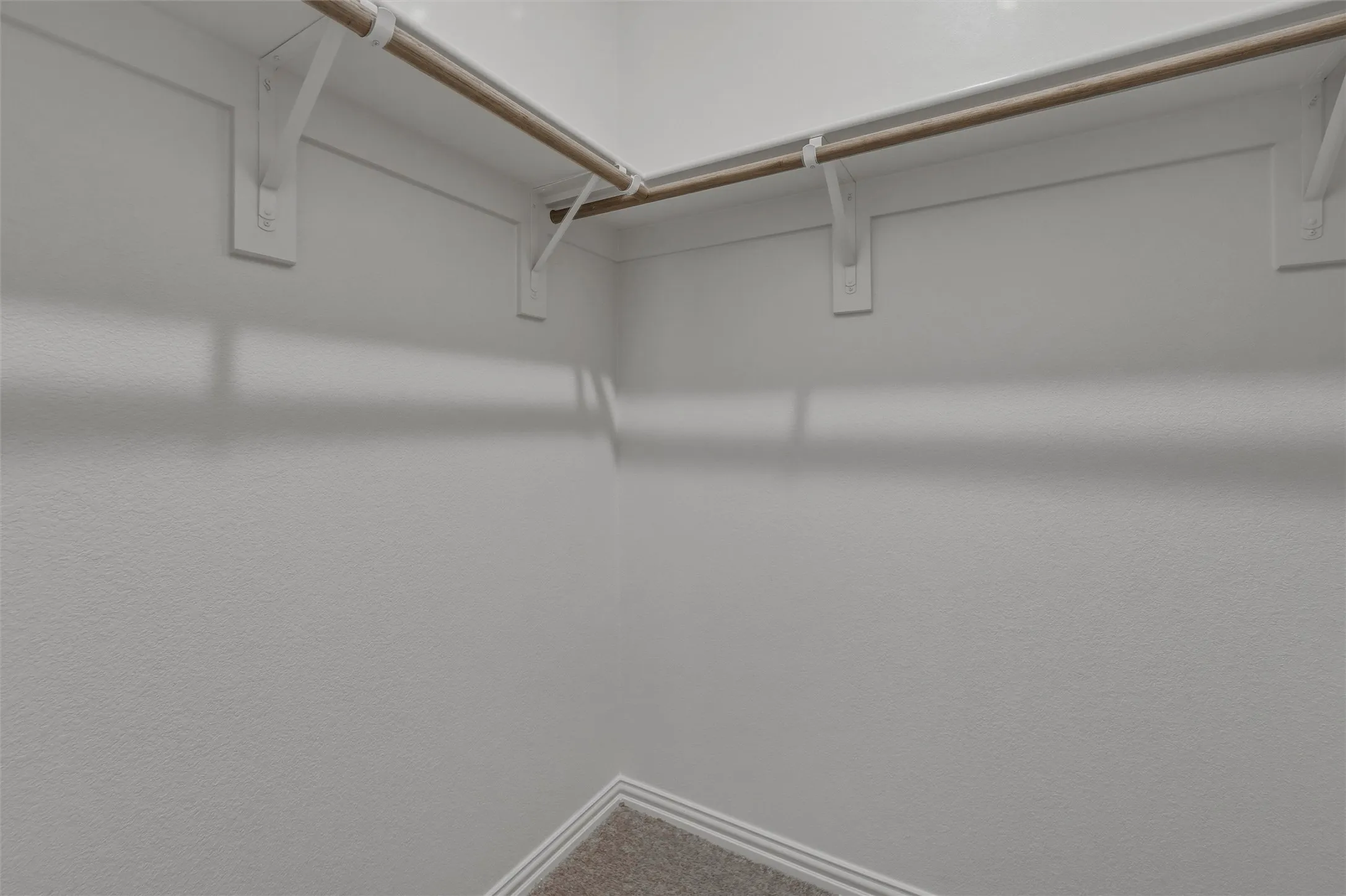 Corner closet space with light neutral wall paint, wood-finish hanging rods, and neutral carpet flooring