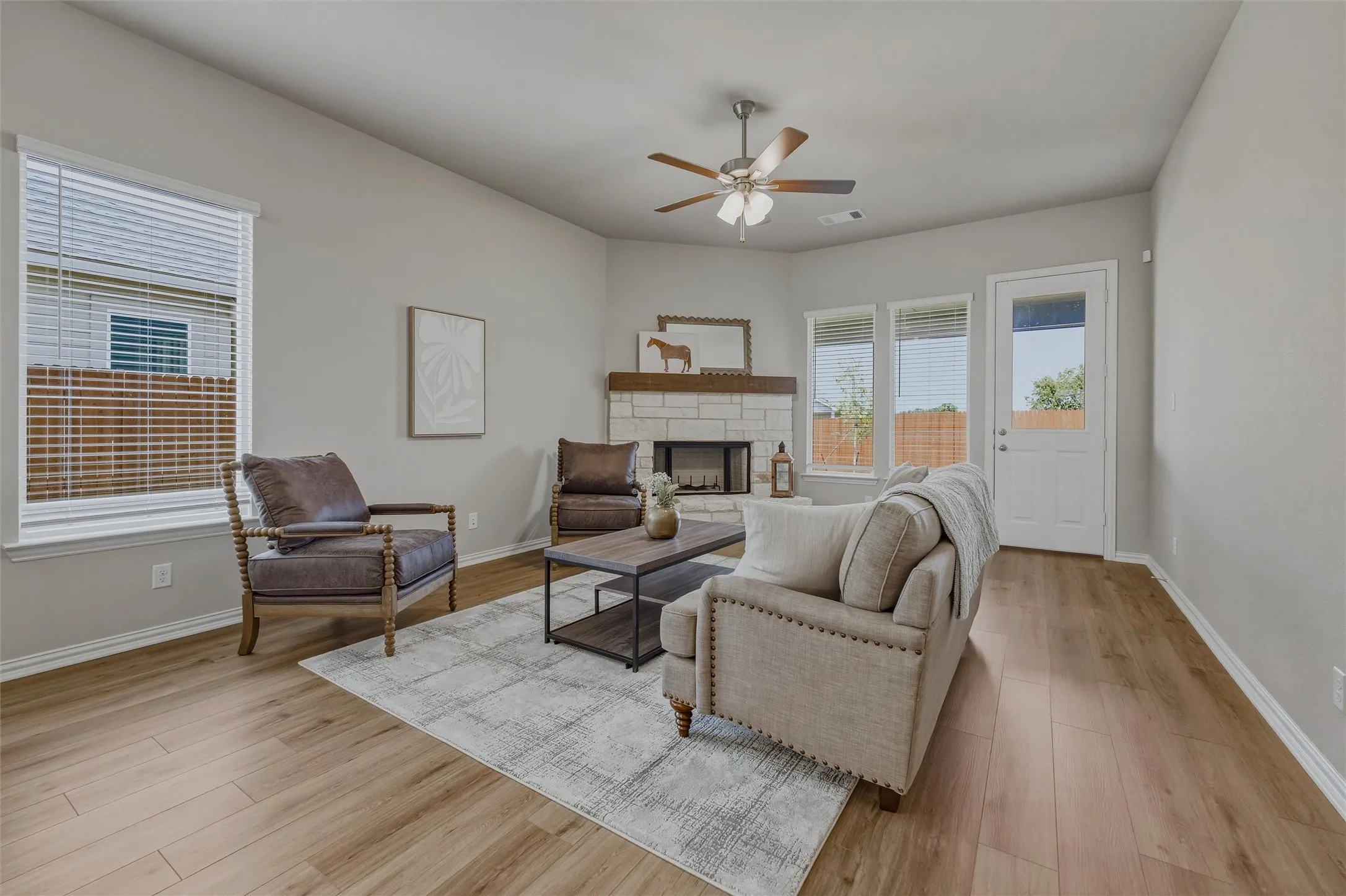 Vaulted ceiling with a ceiling fan, light wood-finish flooring, stone-front fireplace with a wood mantel, and multiple windows with blinds