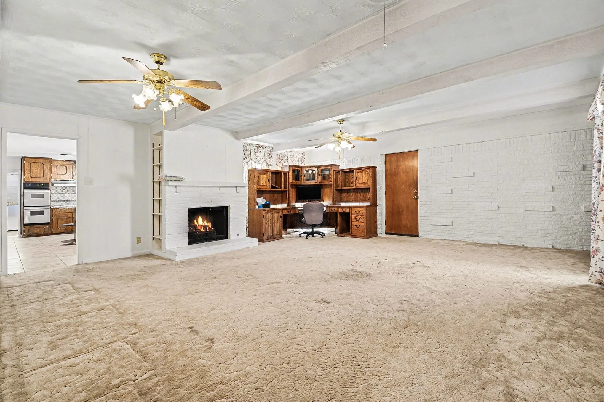 Unfurnished living room with an office area, light colored carpet, a ceiling fan, a brick fireplace, and beam ceiling