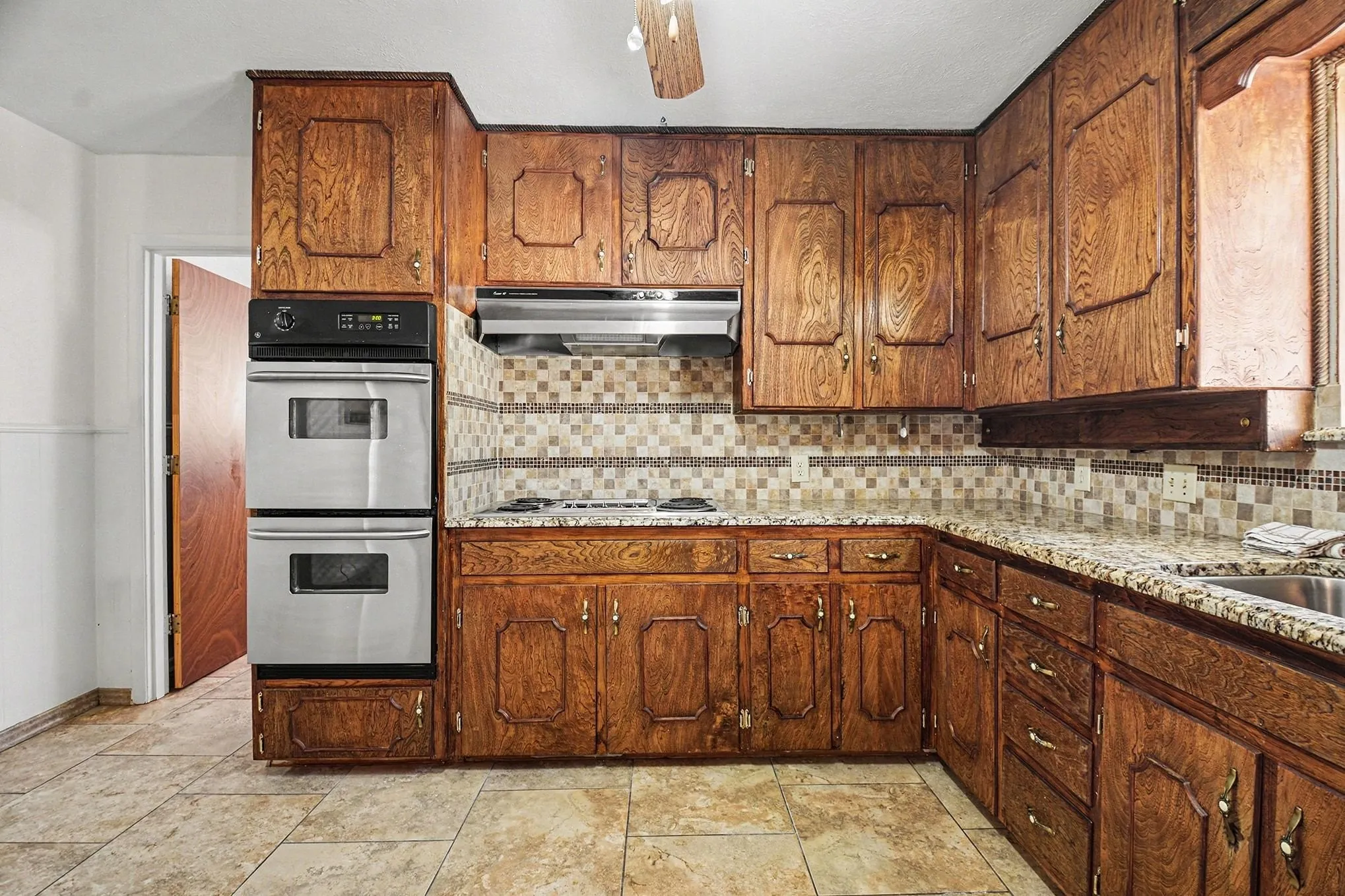 Kitchen featuring stainless steel double oven, tasteful backsplash, light stone counters, wood finish cabinets, and a ceiling fan