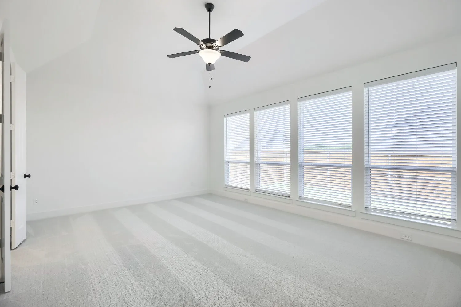 Spacious room featuring vaulted ceilings, a ceiling fan with light fixture, four large windows with horizontal blinds, light-colored carpet flooring, and crisp white walls