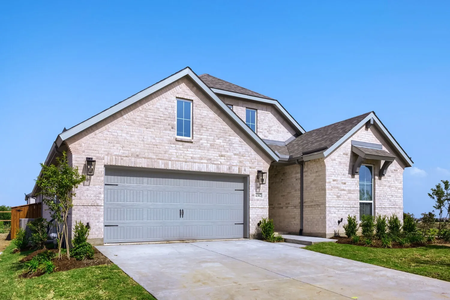 Light brick facade featuring a two-car garage with a panel door, gabled rooflines, decorative exterior lighting, and a prominent arched window with a wood awning