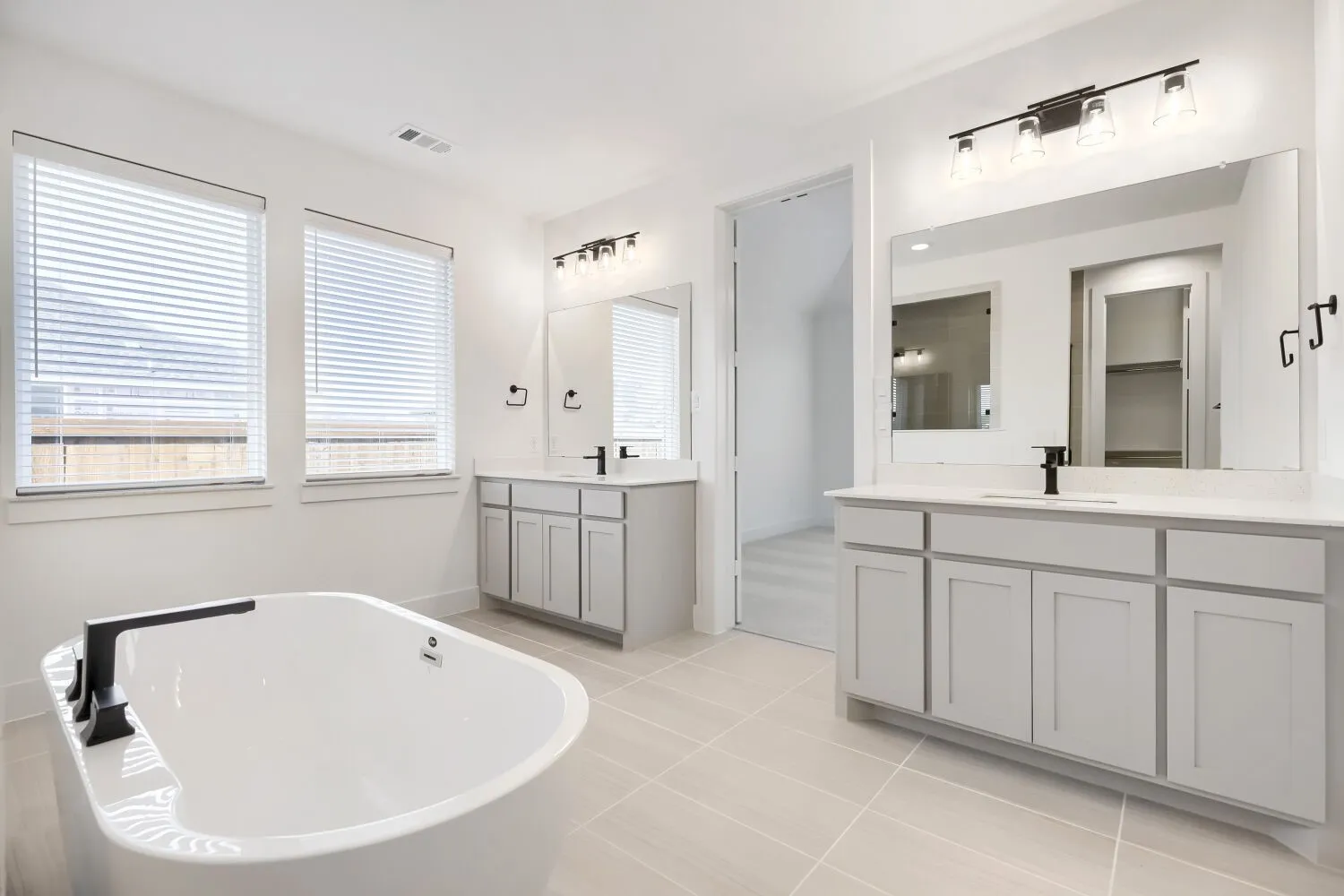 Spacious bathroom featuring a freestanding oval tub with matte black fixtures, dual vanity sinks with light gray cabinetry and white countertops, large mirrors, and tiled flooring