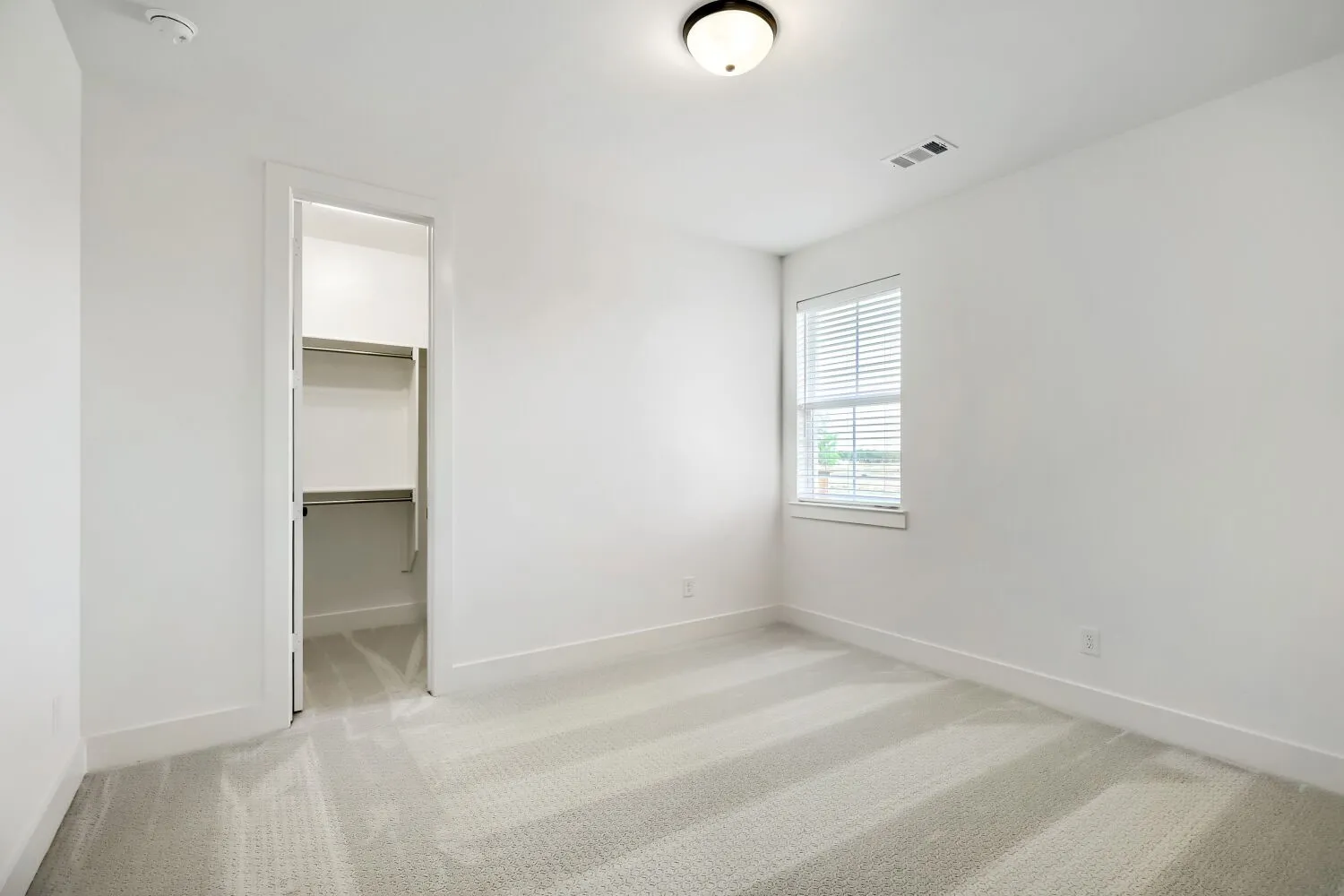 Neutral-toned room featuring a window with horizontal blinds, light-colored patterned carpet, white walls, and a closet with shelving and hanging rods
