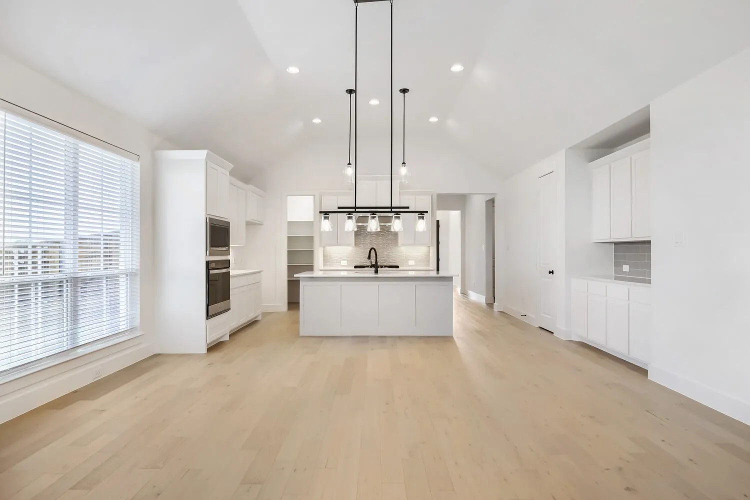 Bright kitchen and living space featuring high vaulted ceilings, light wood-finish flooring, a large kitchen island with a dark faucet, white cabinetry, and built-in double ovens