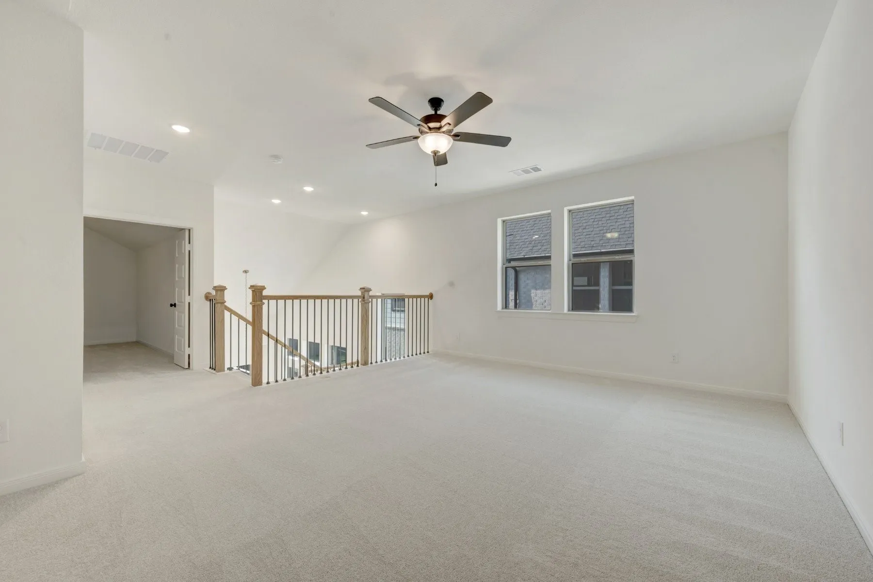 Spacious carpeted room featuring a ceiling fan with integrated lighting, recessed ceiling lights, two windows, and a wood and metal baluster railing overlooking a lower level