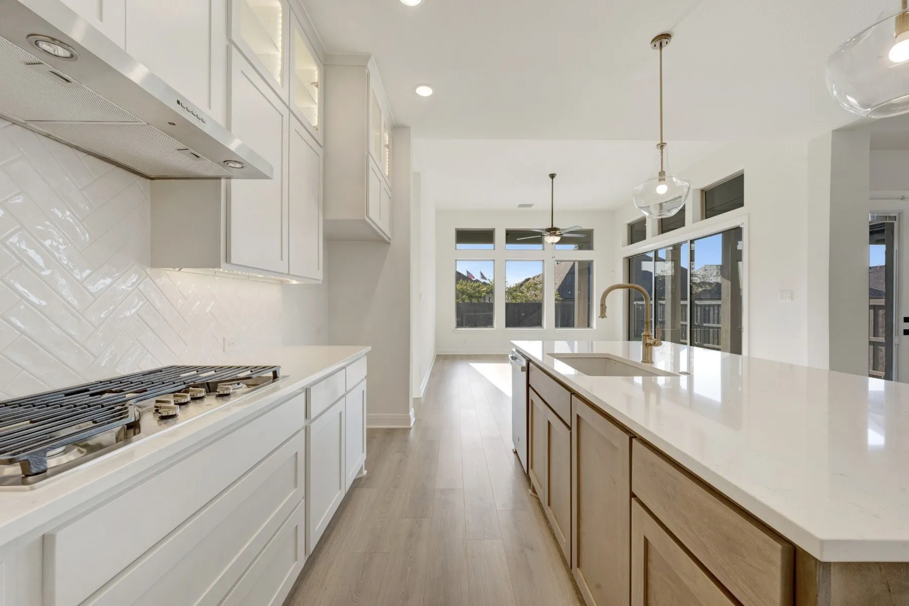 Kitchen featuring an expansive island with an undermount sink and brass-finish faucet, white shaker cabinetry with under-cabinet lighting, a stainless steel gas range with a matching range hood, a white herringbone tile backsplash, and light wood-finish flooring