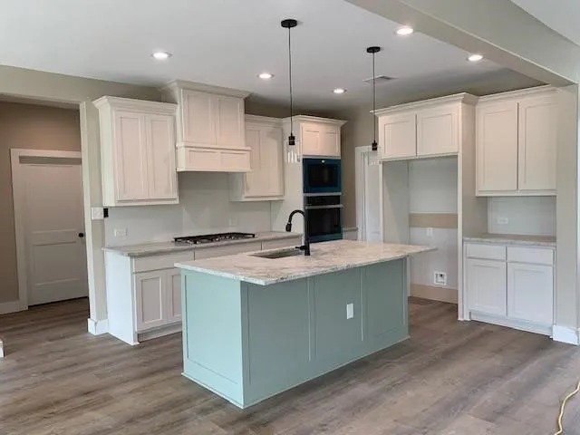Kitchen featuring decorative light fixtures, light wood-type flooring, an island with sink, black appliances, and light stone countertops