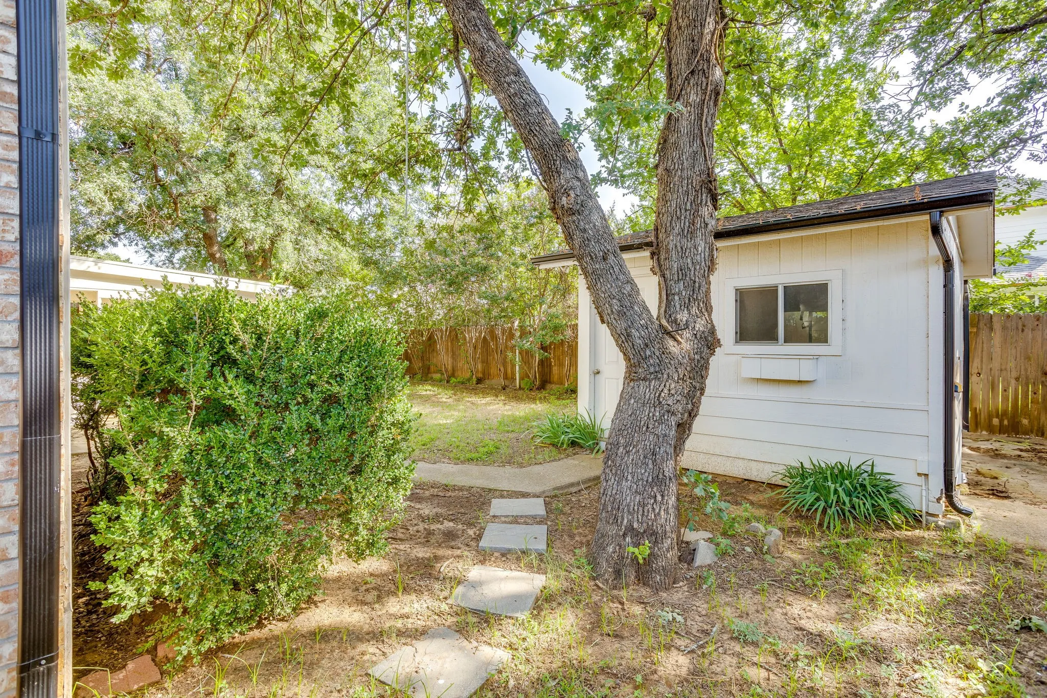 Backyard featuring a detached white outbuilding with a window and window box, mature trees, and concrete stepping stones