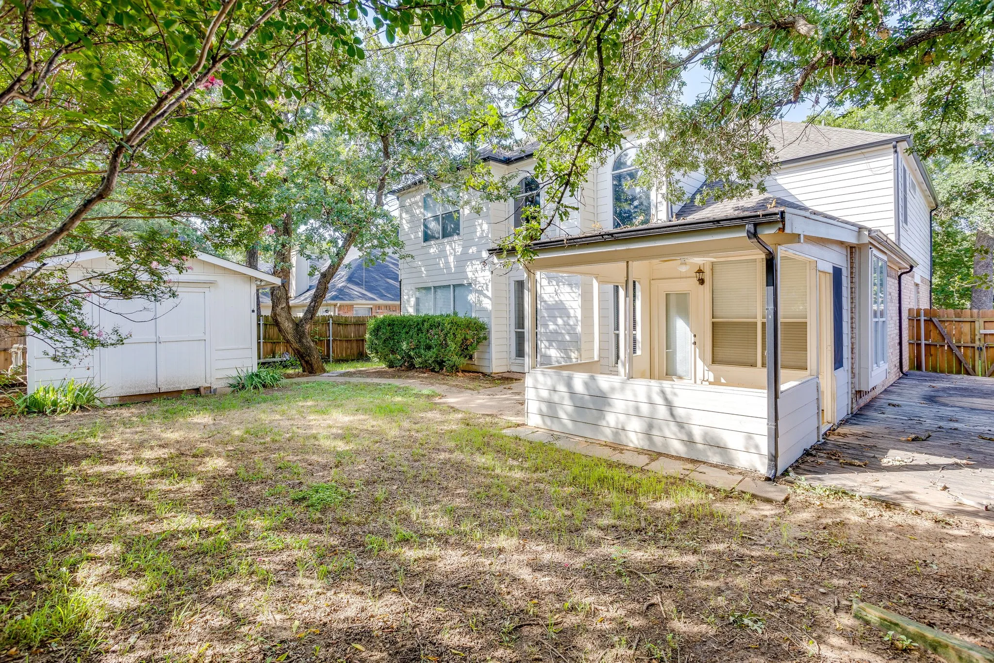 Expansive backyard featuring a white storage shed, mature trees, and a covered patio area with exterior lighting