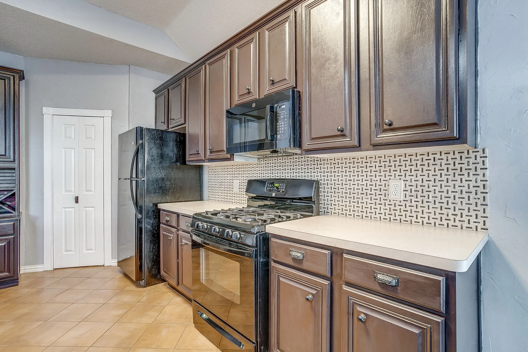 Kitchen with black appliances, decorative backsplash, light countertops, light tile patterned floors, and vaulted ceiling