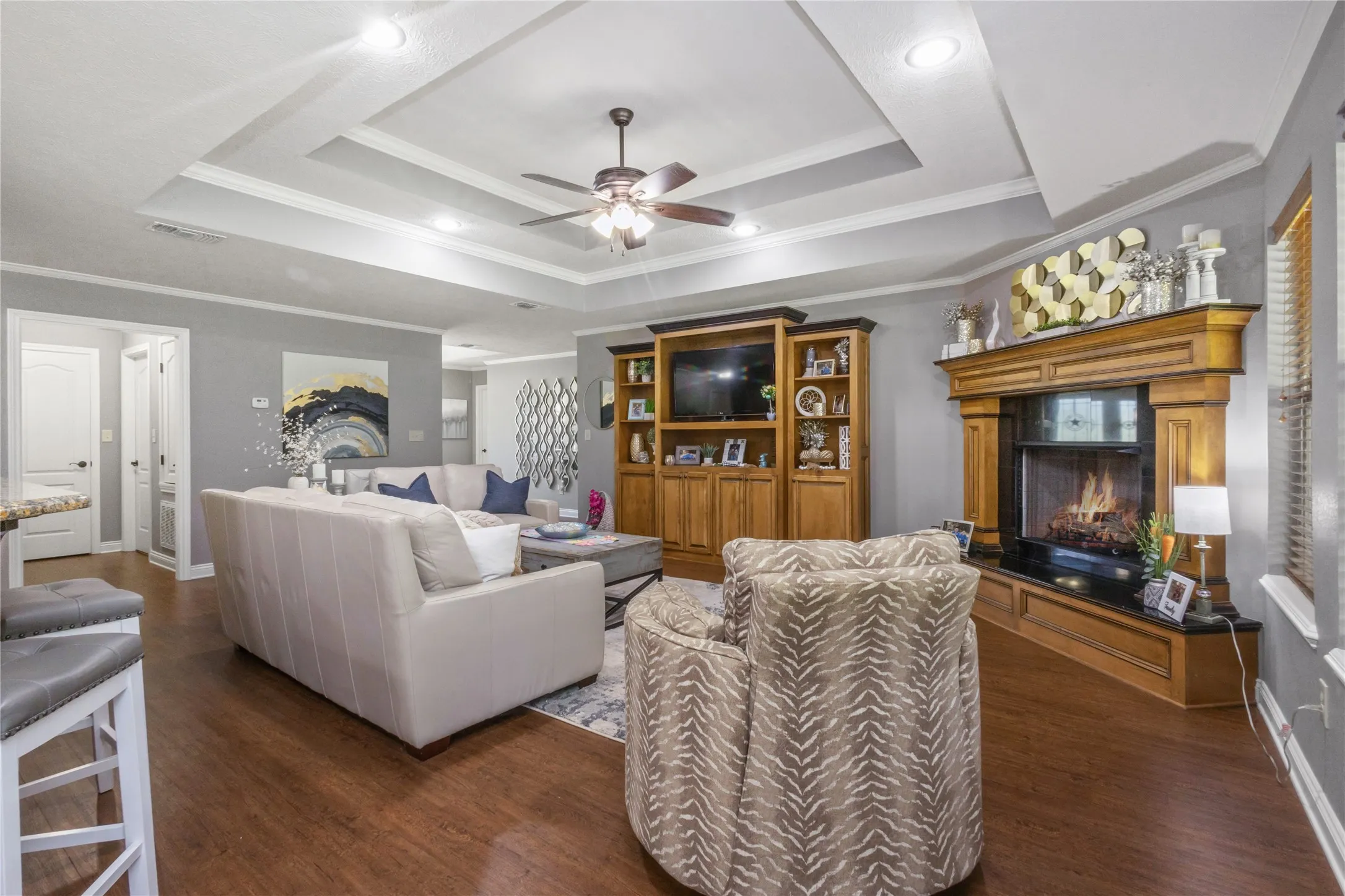 Living room featuring crown molding, a lit fireplace, ceiling fan, dark wood-style floors, and a tray ceiling