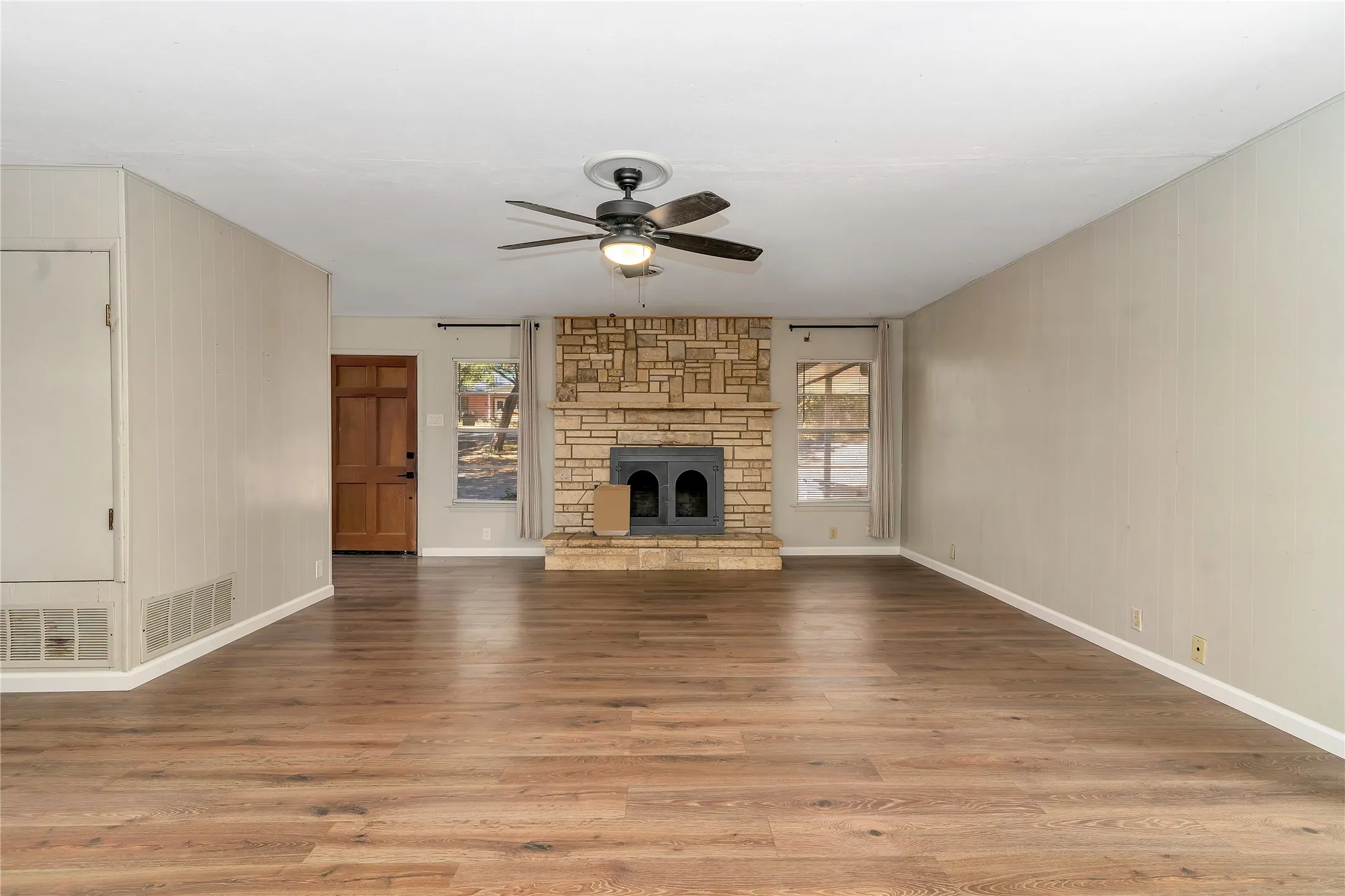 Unfurnished living room featuring a ceiling fan, a stone fireplace, light wood-type flooring, and wooden walls