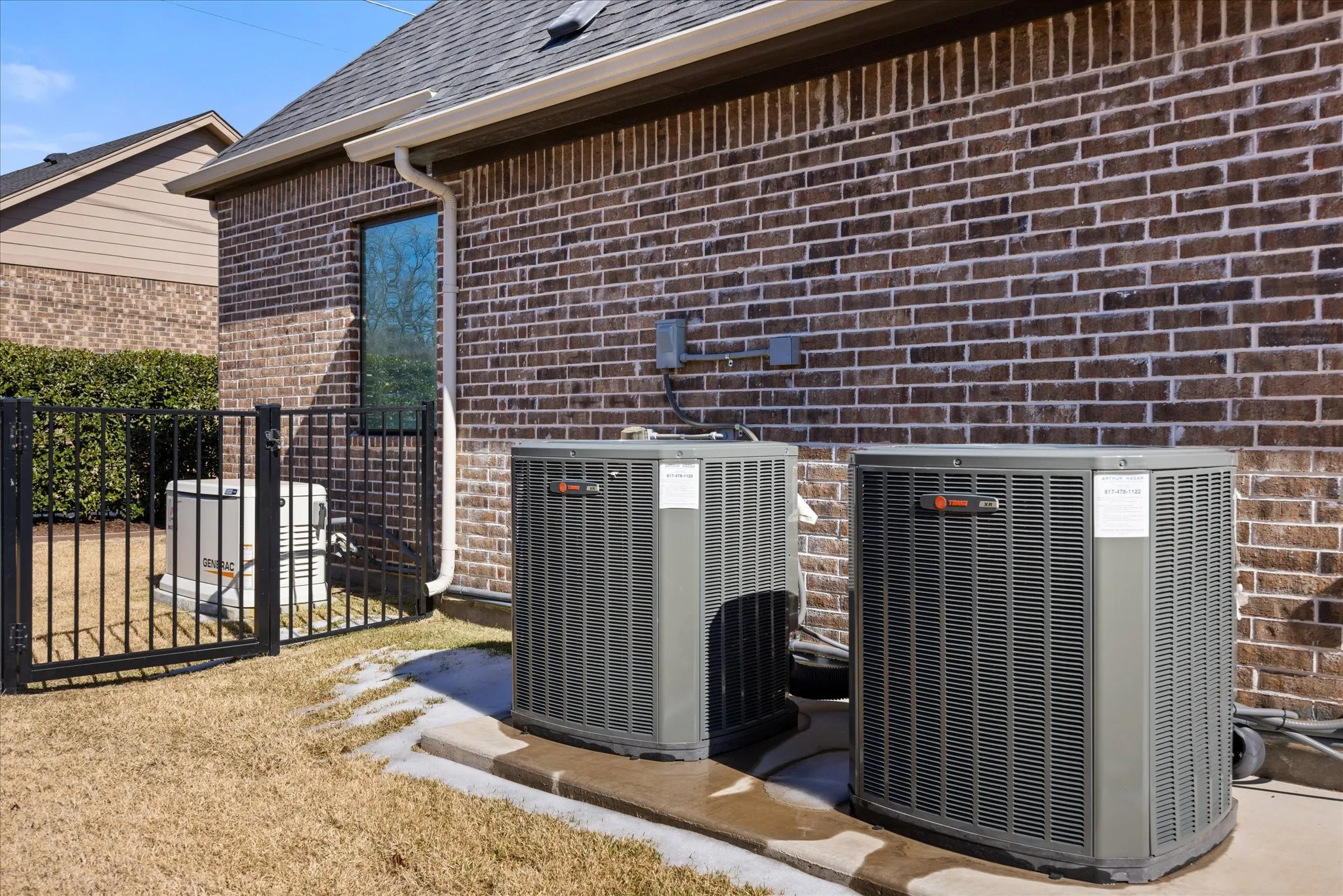 Exterior view of HVAC units and generator, brick siding, roof with shingles, gutters, a downspout, and a gate