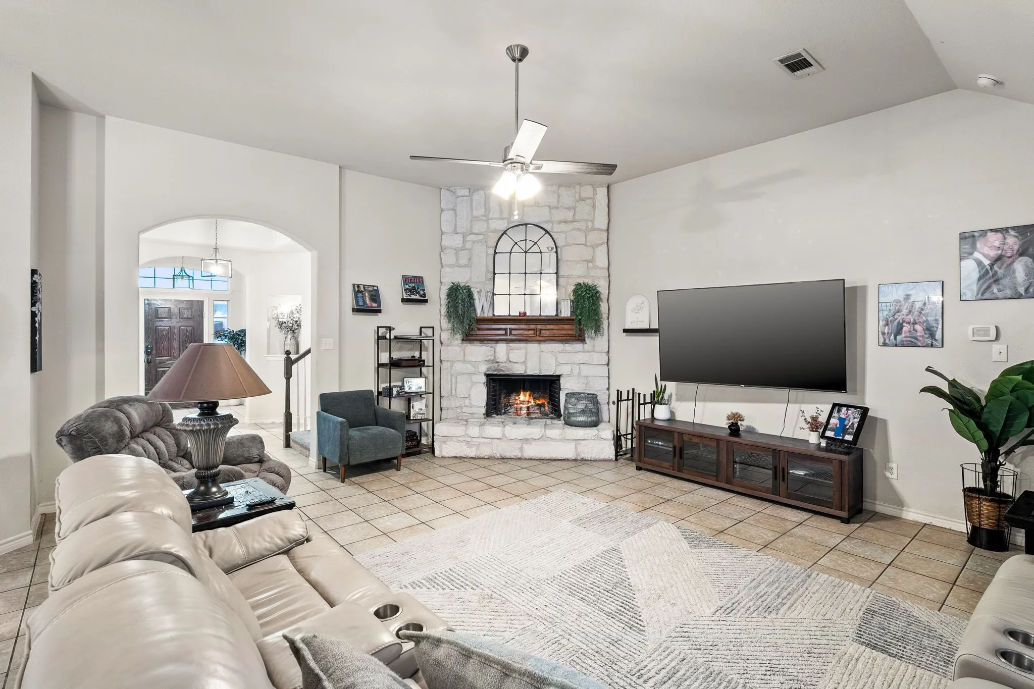 Living area featuring light tile patterned floors, a ceiling fan, a stone fireplace, arched walkways, and vaulted ceiling