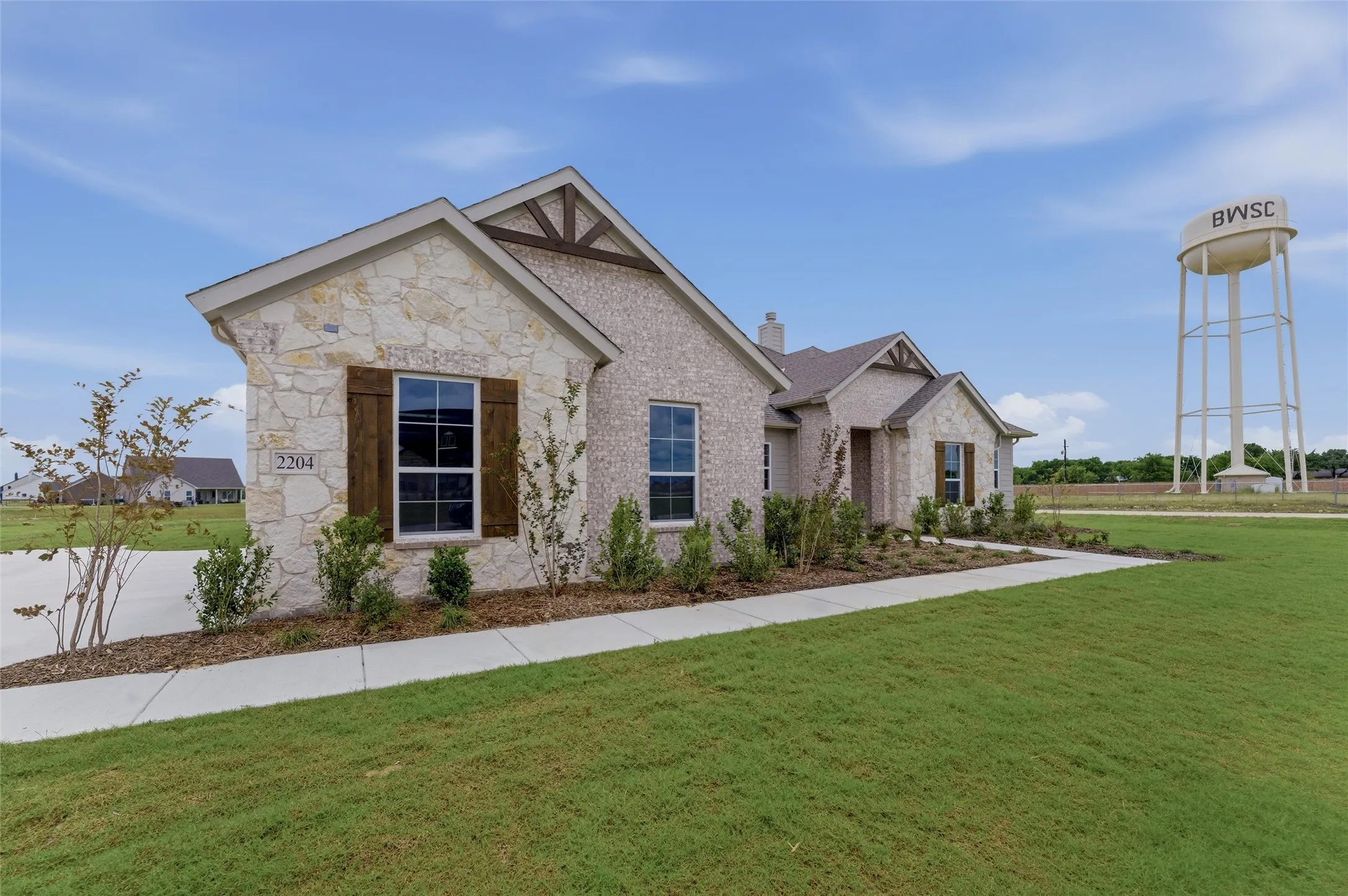 Stone and brick exterior featuring wooden decorative shutters, a concrete walkway, mature landscaping, and an expansive green lawn